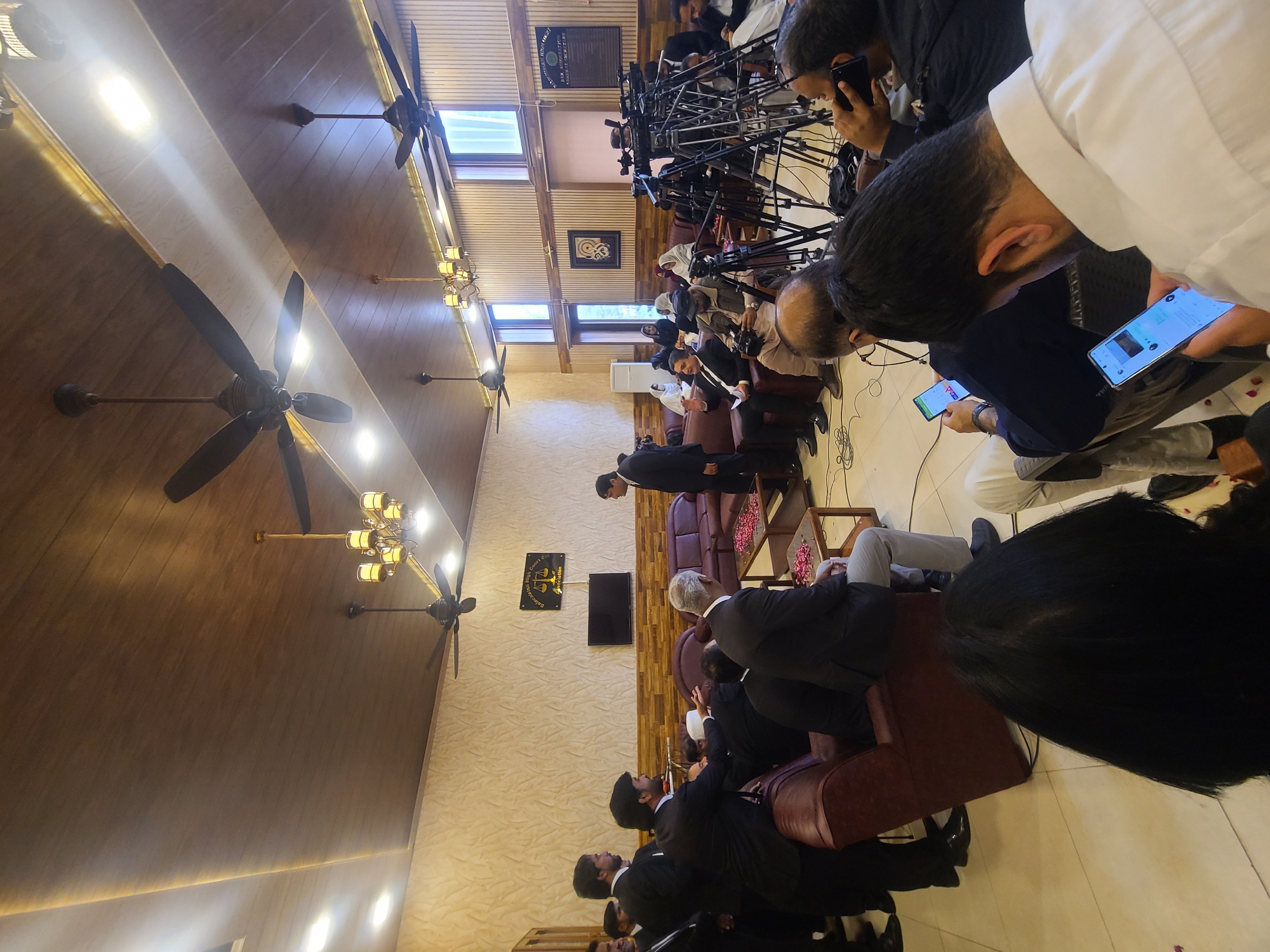 The image shows a gathering of people, some of whom are seated in armchairs and sofas, while others are standing. Many of the people present are wearing dark suits. Several individuals are looking at their mobile phones. A bank of television cameras is positioned at the end of the room. The location appears to be an indoor venue with ceiling fans and chandeliers. The walls are light-colored, with wooden paneling on one section. A sign for the Balochistan High Court Bar Association is visible, indicating the location is likely in Quetta City Tehsil, Pakistan.