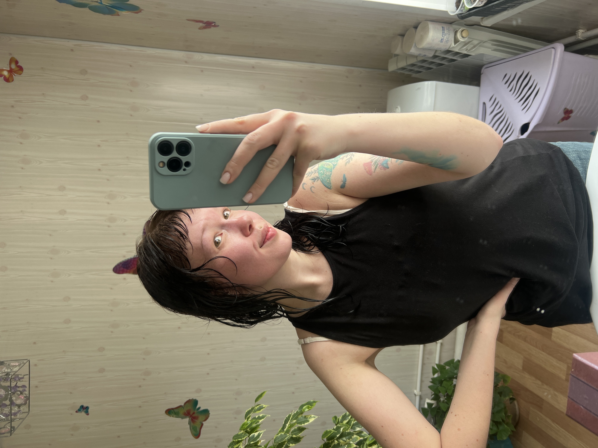 A young woman with wet, dark hair and light eyes is taking a mirror selfie in an indoor setting, likely a bathroom or utility room in Zhigalki, Russia. She holds a light blue/grey smartphone with a triple camera in her right hand, reflecting her image. She has a small gold nose piercing and multiple tattoos on her left arm, including colorful butterflies, a moon, and abstract designs. She is wearing a black tank top, with a light-colored bra strap visible. Her expression is neutral to slightly smiling, suggesting contentment.
The background features a light wooden panel wall on the left, adorned with colorful butterfly decals. A green plant with variegated leaves is partially visible at the bottom left. On the right, a cluttered shelf or counter holds several white cylindrical bottles on a plastic rack. Partial Russian text is visible on one bottle: "...УДЛЯЮЩЕЕ ГЛУБОКО ОЧИЩАЕТ ...ОРОТЬСЯ" (suggesting a deep-cleansing product). A light purple plastic laundry basket or storage bin with vents and a white appliance are also present. The flooring appears to be brown wood. The scene depicts a casual, domestic environment during daytime.