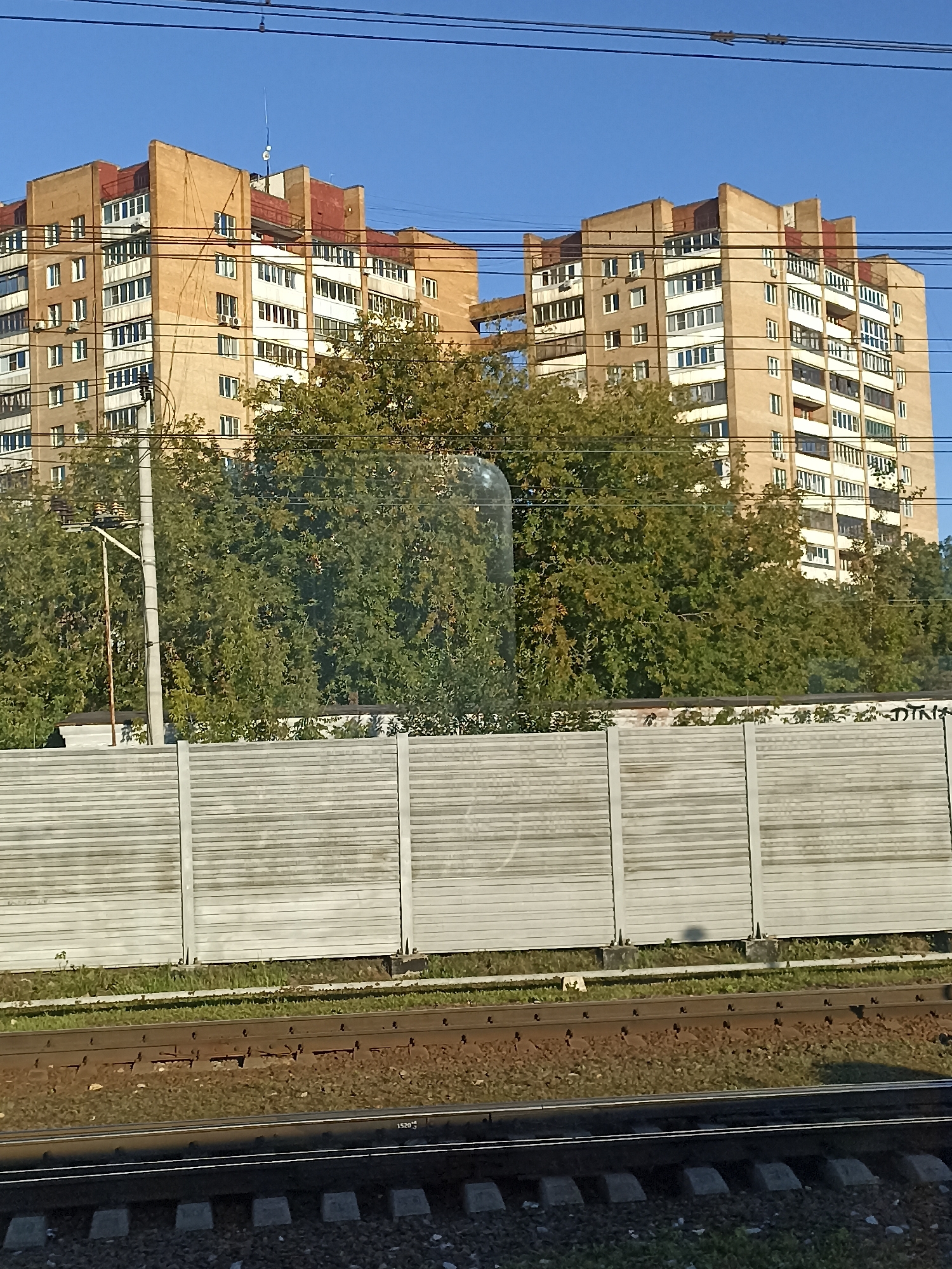 The image shows a view of a residential building and its surroundings in Podolsk, Russia. Two apartment buildings, appearing to be constructed of beige brick and white sections, dominate the upper portion of the frame, with a clear blue sky above. They feature multiple floors with numerous windows and balconies. Lush green trees with some signs of autumnal color are situated between the buildings and a concrete barrier. This barrier, constructed of horizontal, ribbed panels, runs across the middle ground of the image. In the foreground, railway tracks are visible, with gravel ballast and concrete sleepers. A utility pole with wires is positioned to the left, partially obscured by the trees. The scene appears to be captured on a sunny day.