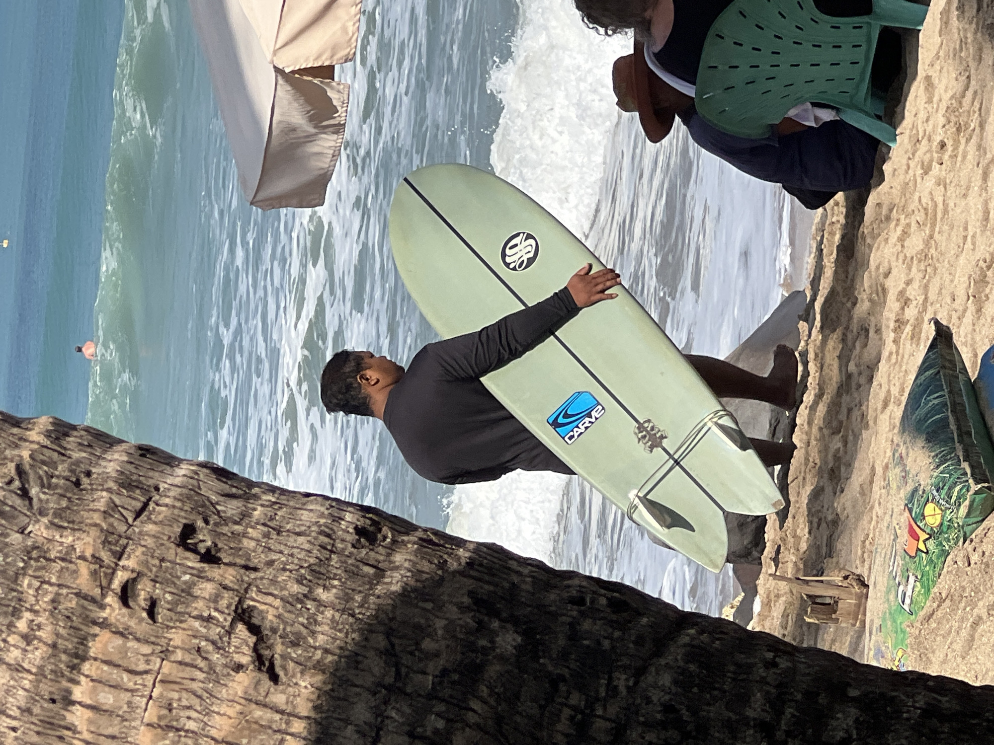 In Kuta, Indonesia, on a sandy beach, a man in a black long-sleeved shirt stands near the water's edge, holding a light green surfboard. The surfboard has a black outline and two black stripes running down its length, with a circular logo on the left side. A blue "CARVE" logo is visible on the lower portion of the surfboard. The man is facing away from the camera, towards the ocean. In the background, the turquoise ocean has waves breaking, and a person is visible swimming in the distance. To the right, part of a cream-colored beach umbrella is visible. In the foreground, to the left, a portion of a large palm tree trunk is in shadow. On the sand, to the right of the man, two people are seated, one wearing a brown hat and the other wearing dark clothing. A green plastic chair is also present.