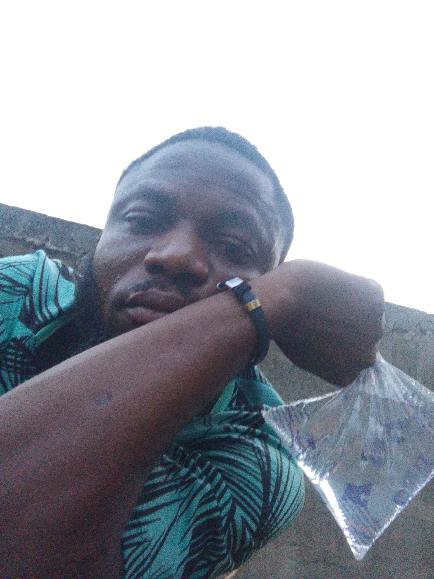 A man is positioned outdoors, holding a plastic bag of liquid in his right hand. He rests his head on his left fist, which is adorned with a watch. The background includes a gray wall and sky. The man wears a shirt with a teal and black leaf pattern. The location is Lokoja, Nigeria.