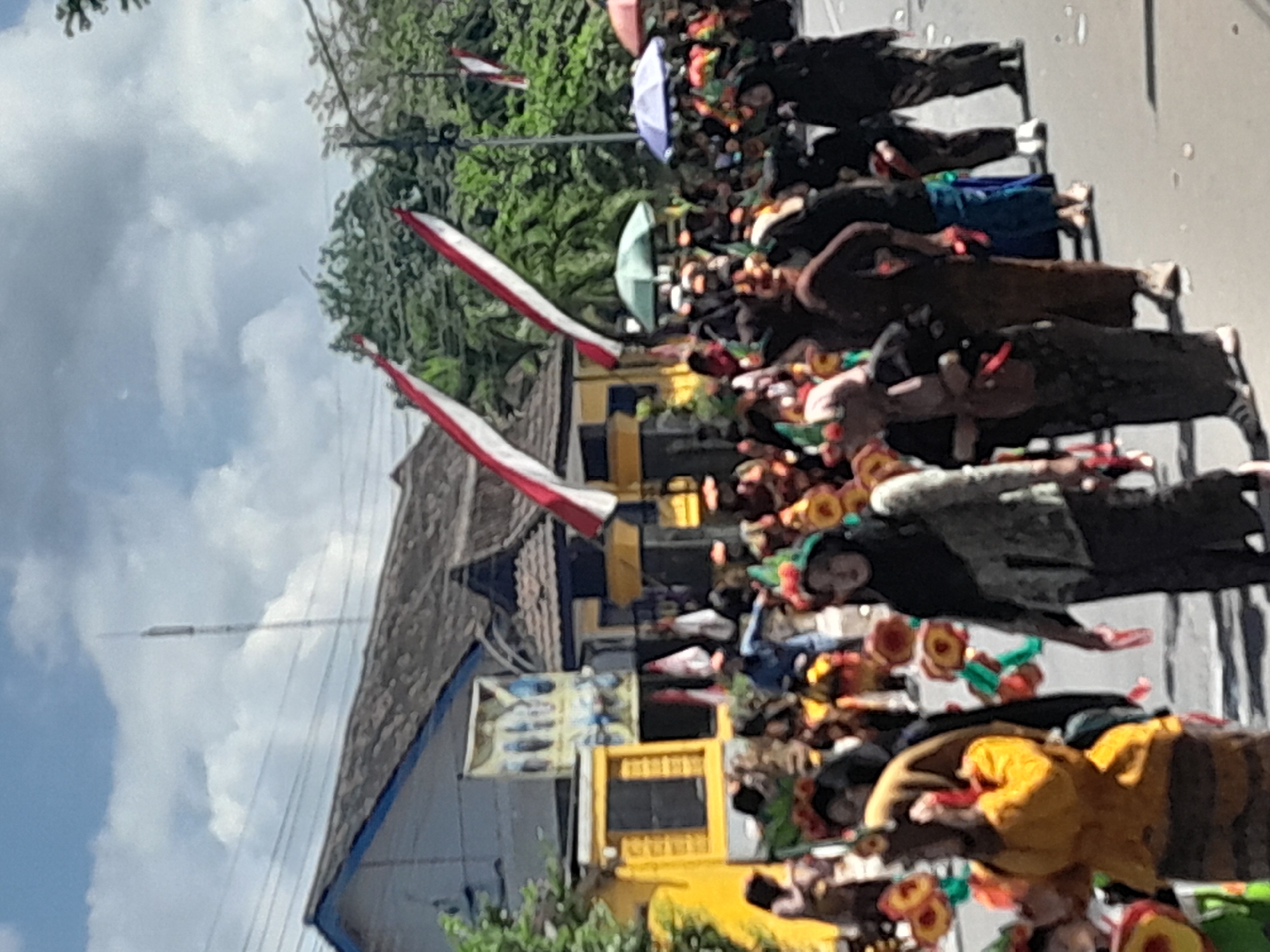 The image depicts a procession in Genteng Kulon, Indonesia. A group of people, many wearing traditional Indonesian attire and headdresses, are walking along a paved street. Some are holding decorative items that resemble flowers. In the background, there are buildings with distinctive tiled roofs, one of which is painted yellow and features a banner. Red and white flags, likely the Indonesian flag, are visible in several locations. The sky is partly cloudy. The scene appears to be a cultural event or parade.