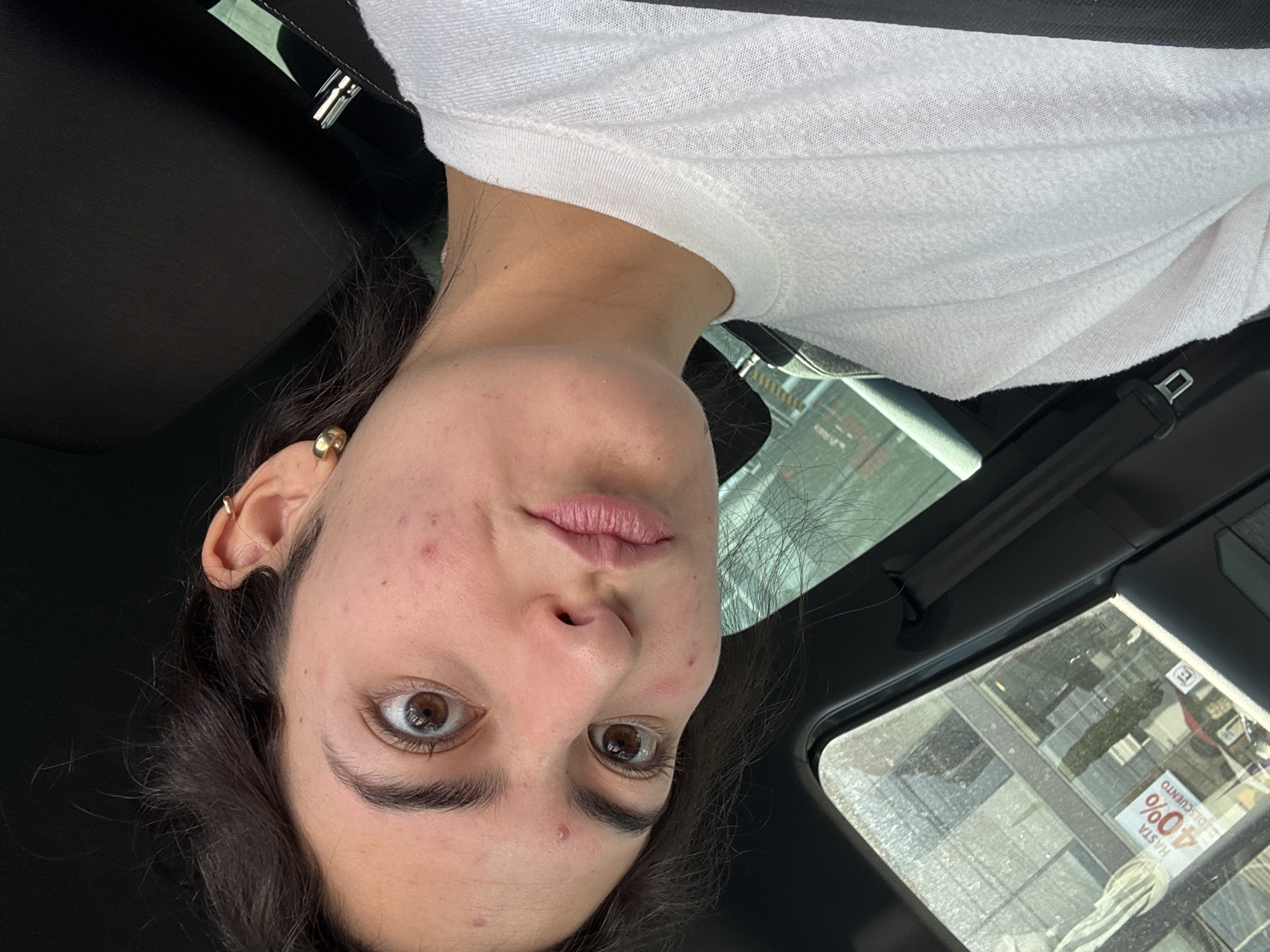 The image captures a young woman, likely in her early twenties, inside a car. She has dark brown hair, wide brown eyes, and light-to-medium skin tone with some visible blemishes on her forehead and cheeks. She is wearing a simple white short-sleeved t-shirt and has gold hoop earrings in both ears. Her expression is neutral to slightly pensive, with her lips slightly parted.
The setting appears to be the interior of a vehicle, possibly from the passenger seat, with dark upholstery visible around her and a seatbelt buckle in the background. It is daytime, with light filtering through the car windows. Through the window on the right side of the frame, reflections of urban buildings and an advertisement are visible. The advertisement clearly displays the Spanish text "HASTA 40% DE DESCUENTO" (Up to 40% off). The overall scene suggests a candid moment in an urban environment, consistent with the location context of Puebla, Mexico.