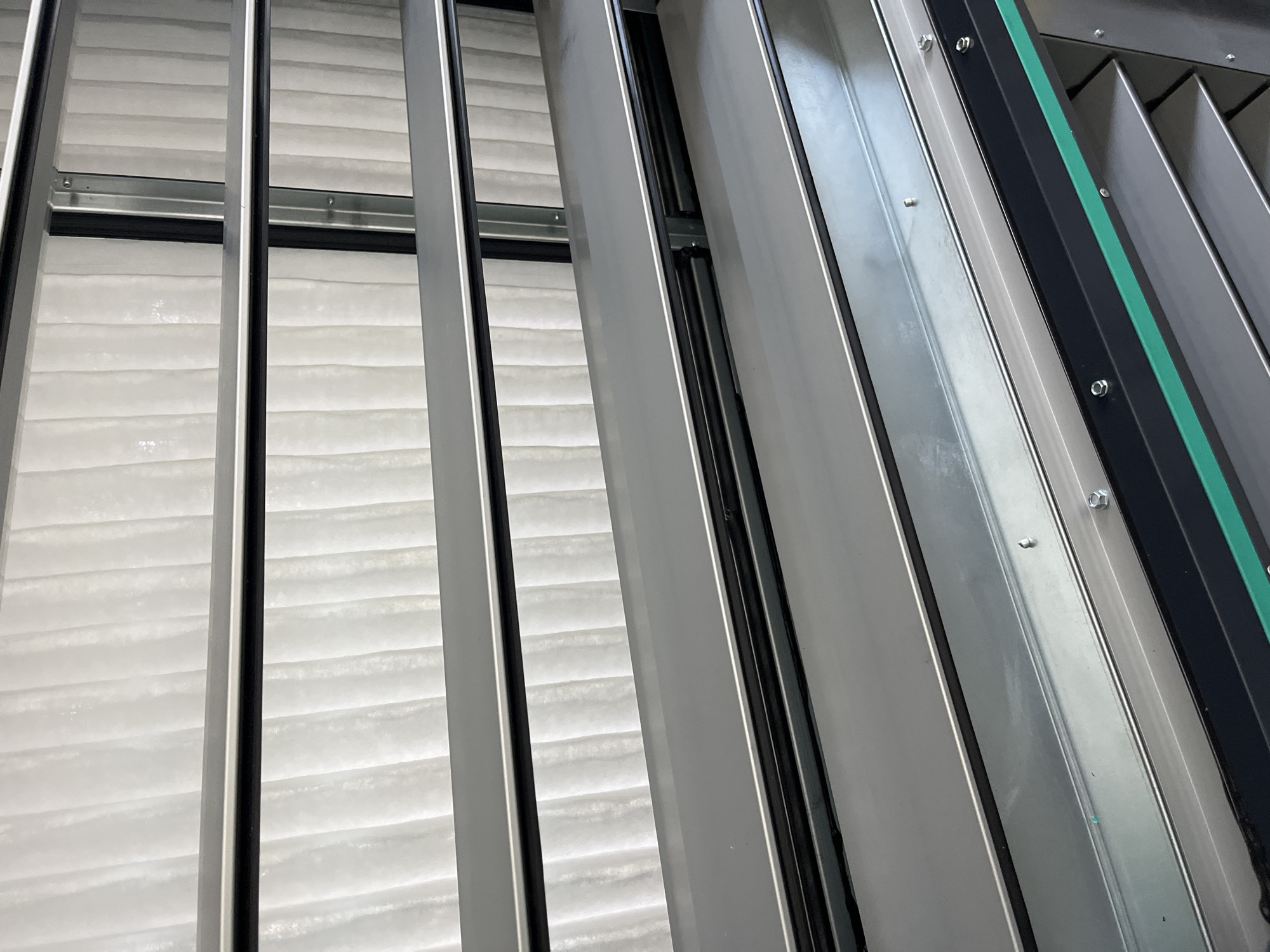 The image shows a close-up view of industrial louvers or vents, likely part of a ventilation system. These louvers are made of gray metal and are stacked horizontally. Behind the louvers, a white, textured material is visible, possibly insulation or filter material. The overall impression is of a utilitarian structure, possibly located in an industrial area or a large building. The image does not provide any specific details to determine its exact location within Kyiv, Ukraine, or any context about people, actions, or interactions.