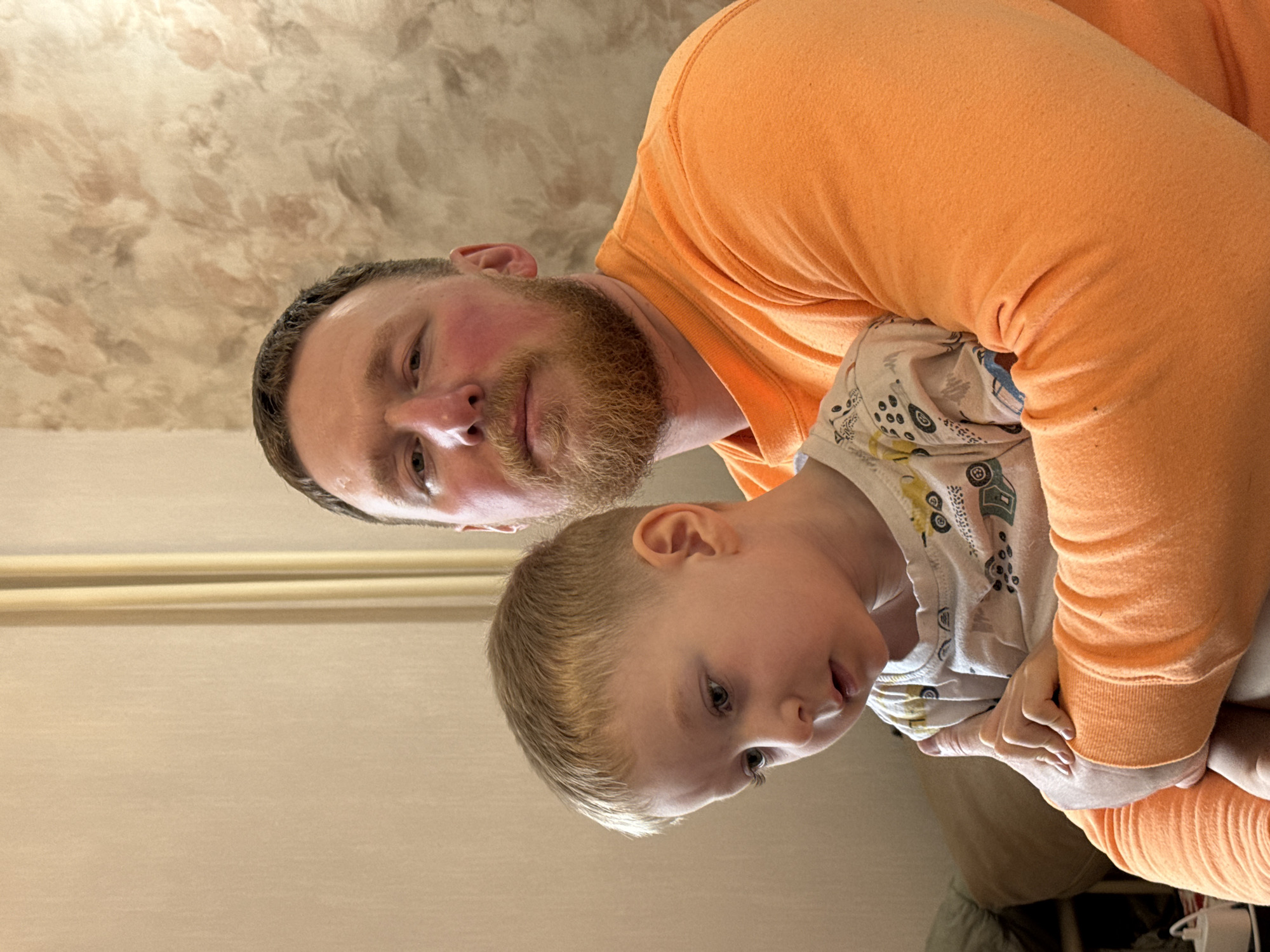 A man with a reddish beard, wearing an orange long-sleeved shirt, is holding a young child in his arms. The child is wearing a light-colored, long-sleeved shirt with a pattern of vehicles. The photograph is taken at an odd angle, seemingly looking up at the subjects. The background appears to be a room with light-colored walls. There is no information to place this image in the city of Perm, Russia.