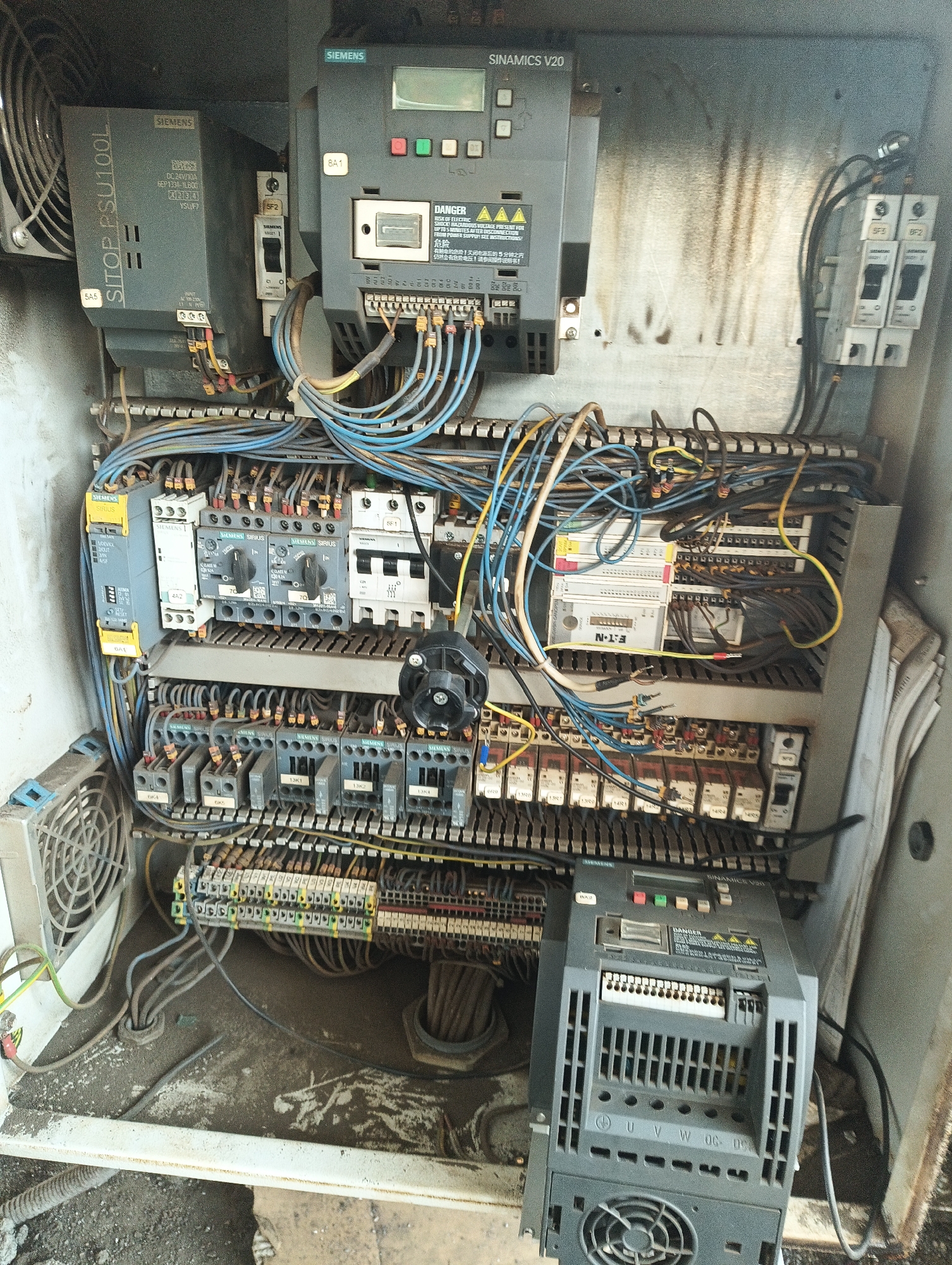 The image shows an open electrical control panel with various Siemens components. At the top left is a Siemens SITOP PSU100L power supply unit, along with circuit breakers labeled 5F2 and SF2. A Siemens SINAMICS V20 drive is also present, featuring a warning label with electrical hazard symbols and text in English and Chinese. Below these are rows of Siemens Sirius contactors and relays. An Eaton N-1 component is visible towards the right side of the panel. The panel is filled with numerous wires and connectors. The general environment suggests an industrial setting. The location is Unknown, Unknown.