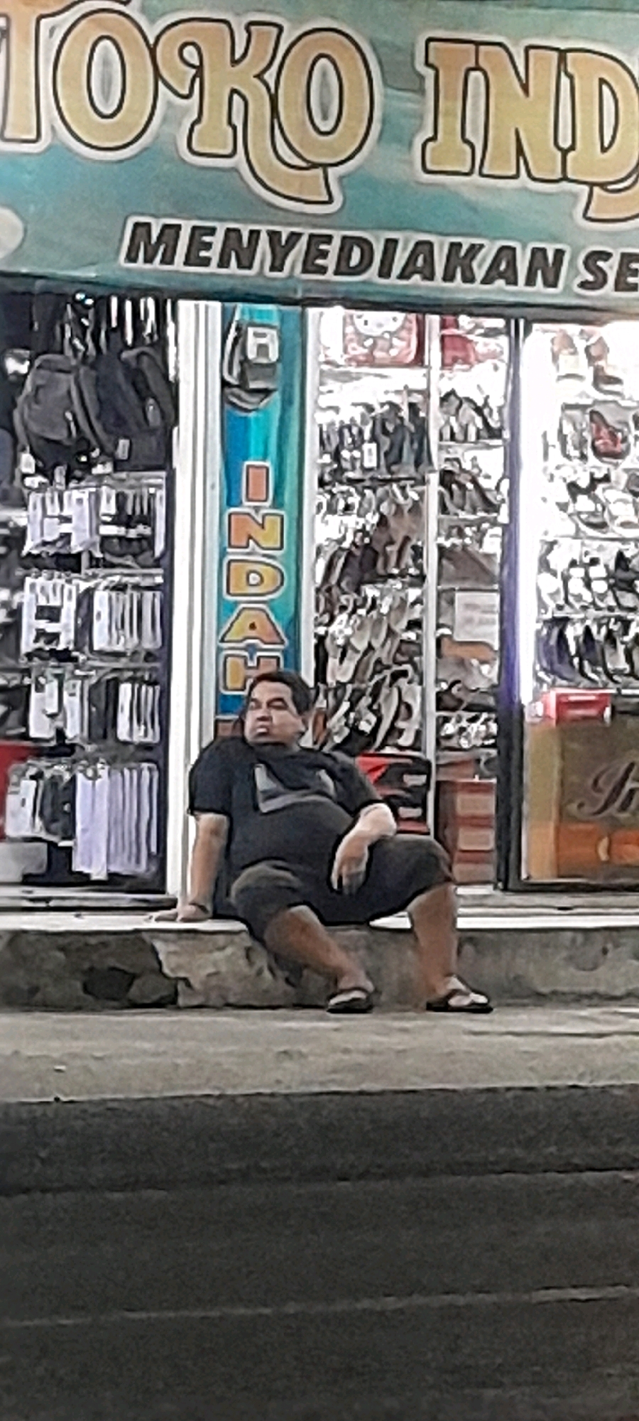 A man is sitting on a concrete step in front of a store in Rogojampi, Indonesia. The store has a sign that reads "TOKO INDAH." The man is wearing a black t-shirt and dark shorts. He is looking down and to his left. In the background, the store displays various items, including bags, shoes, and clothing. The overall scene appears to be outdoors, possibly on a street or sidewalk.