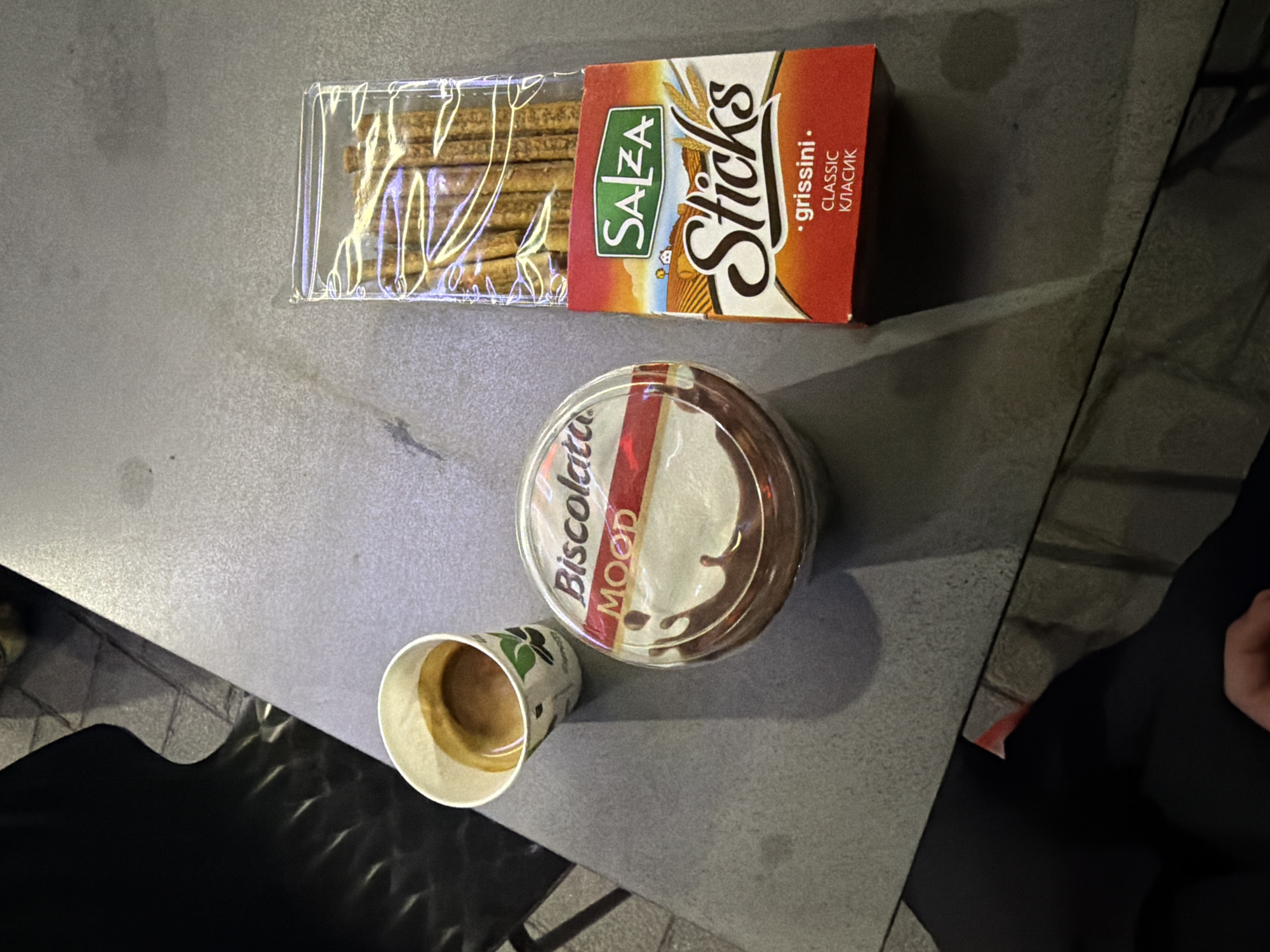 The image displays a table with a cup of espresso, a container of "Biscottata Mood" chocolate spread, and a package of "Salza Sticks" grissini. The objects are arranged on a textured surface, possibly a table in an outdoor seating area. The lighting suggests it is daytime, with shadows indicating sunlight. The background is blurred, showing what appears to be paving stones and dark, indistinct shapes. There are no people clearly visible in the frame, although a hand is partially visible in the lower right corner. The scene is forensic and matter-of-fact, documenting the presence and arrangement of these food items. The location of Zvornik, Bosnia and Herzegovina, is not discernible from the visual information provided in the image.