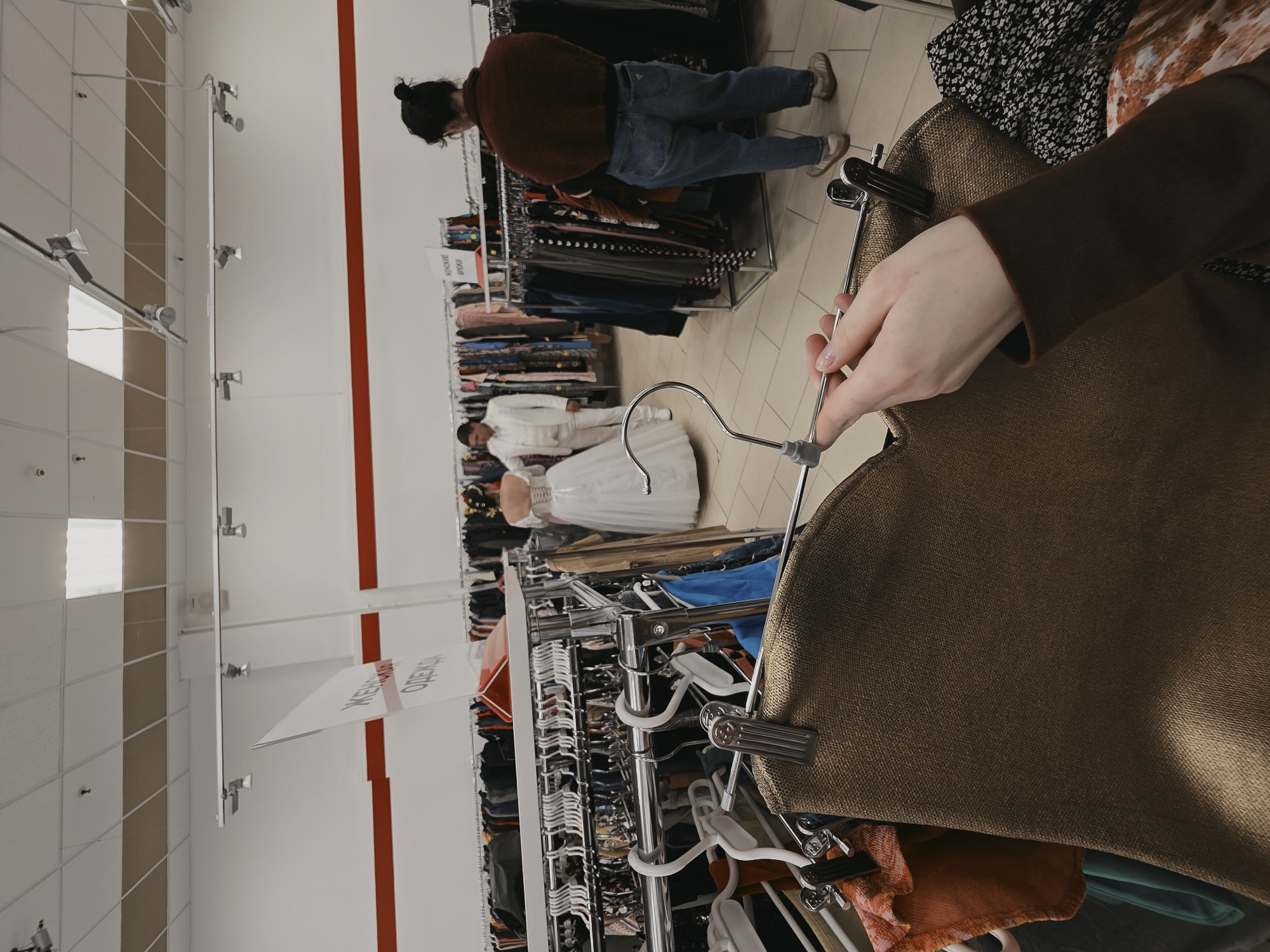 The image shows the interior of a clothing store, specifically the section for women's clothing as indicated by the sign “ЖЕНСКАЯ ОДЕЖДА”. Clothing racks are visible, densely packed with items. A person is holding a hanger with a brown garment, likely a coat or jacket. In the background, two figures can be seen: one wearing a white suit and the other wearing a white dress. Another person is browsing through a rack of clothing. The location is within the city of Saint Petersburg, Russia, as inferred from the signage in Russian.