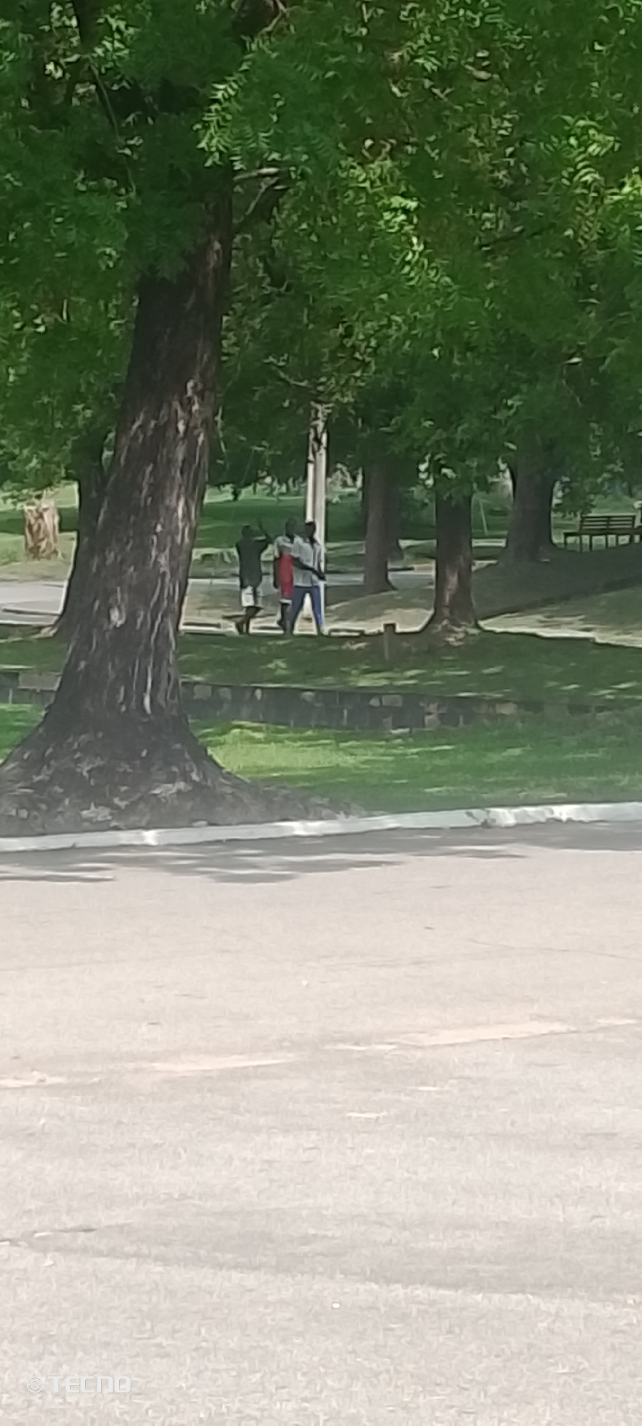 The image captures a group of three individuals standing outdoors in a park-like setting. Two of the individuals are in the foreground, appearing to be engaged in conversation or interaction. The third individual is slightly behind them, also standing. The scene is characterized by lush green trees with dense foliage, suggesting a warm, possibly sunny day with good weather. In the background, there are more trees and a hint of a pathway or road. The ground appears to be covered in grass and some paved areas. A park bench is visible in the far background. The overall atmosphere seems relaxed and casual. There are no clear indicators of the time of day beyond the general brightness suggesting daylight. There is a visible watermark in the bottom right corner of the image that says "TECNO". The location is identified as Zango-ndaji, Nigeria.