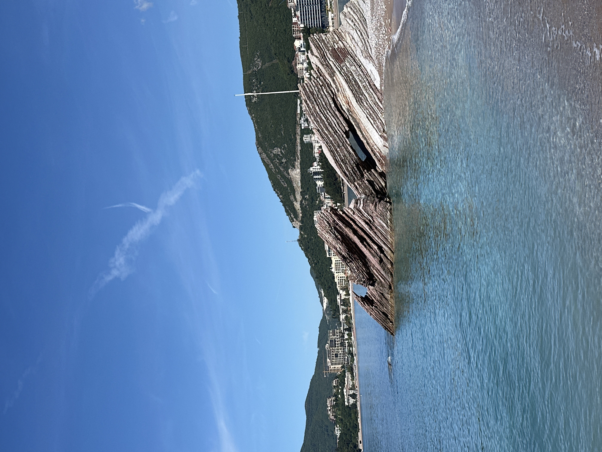 The image presents a coastal scene, likely captured in the vicinity of Boreti, Montenegro, judging by the coastal landscape and architecture.
Environment:
The sky is clear with a light contrail. A coastal mountain range with scattered buildings runs along the shore. The sea is a gradient of light blue and grayish-blue, indicating varying depths or the presence of a sandy beach.
Objects:
A prominent rock formation with visible strata juts out into the sea. Coastal buildings and vegetation populate the mountain slopes. A white structure, possibly a flagpole, is visible on the mountain.
Actions/Interactions:
There are no discernible human actions or interactions visible in the frame. The waves are gently lapping on the shore.