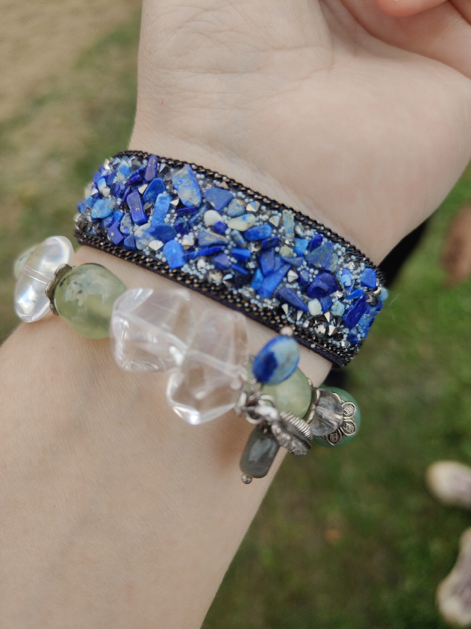 This image shows a close-up view of a person's wrist adorned with two bracelets. The top bracelet is wide and embellished with numerous small, irregular pieces of blue lapis lazuli and glittering accents, bordered by a dark band. Below it, a second bracelet is composed of several larger beads, including clear, faceted crystals, milky white oval stones, and a translucent green bead. Dangles of smaller beads, charms, and a dark grey stone hang from this lower bracelet. The background consists of softly blurred green grass and a light brown, earthy area. The location is not determinable from the visual information.