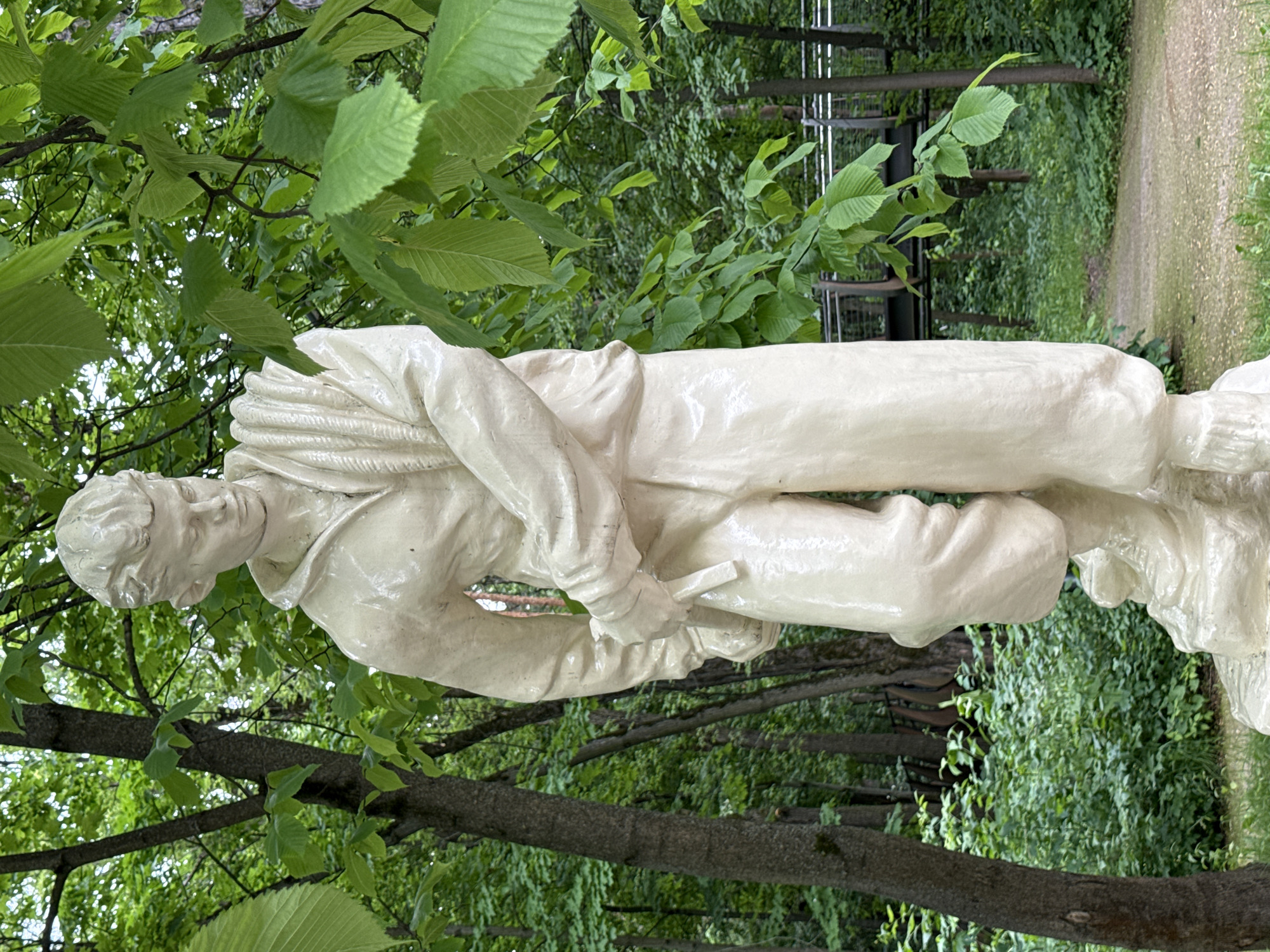 A full-body, light-colored statue of a young man stands in a park on a cloudy day. The statue is made of a glossy material, possibly painted plaster or ceramic, and depicts the man with his left arm across his chest and his right hand holding a sash or belt. He is wearing loose-fitting pants and a jacket with a collar, and his hair is styled in a quiff. The statue stands on a pedestal that blends into the ground, which appears to be covered in dirt and grass.
The background of the image shows a lush green park with mature trees. In the distance, a modern, elevated pedestrian walkway with metal railings can be seen winding through the trees. The foliage is dense, with vibrant green leaves dominating the scene. The overall impression is one of a public art installation set within a natural environment. This statue is located in the Gorky Park (Park of Culture and Leisure named after Gorky) in Moscow, Russia.