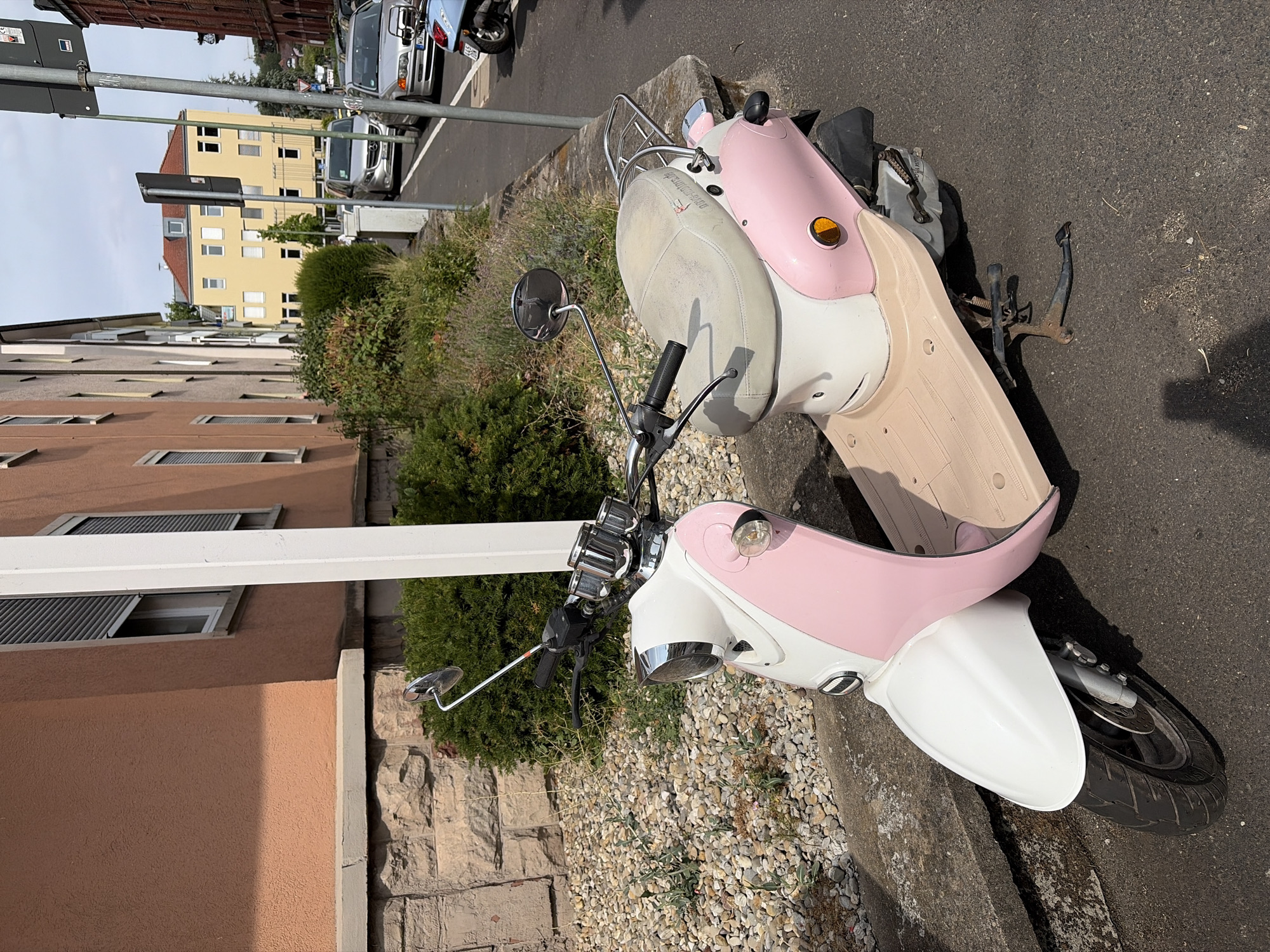 A two-tone pink and white scooter with a light tan floorboard is parked on an asphalt street next to a low concrete retaining wall. The scooter has chrome accents and a single rearview mirror. Behind the scooter, a textured, light brown building with gray-shuttered windows rises. Adjacent to this is a section of rough-hewn stone wall and dense green shrubbery. Further down the street, other vehicles, including cars and another scooter, are visible, along with buildings of various colors and architectural styles, suggesting an urban environment. The sky above is overcast. This scene appears to be on Theaterstraße in Würzburg, Germany, a street known for its mix of residential buildings and commercial activity.