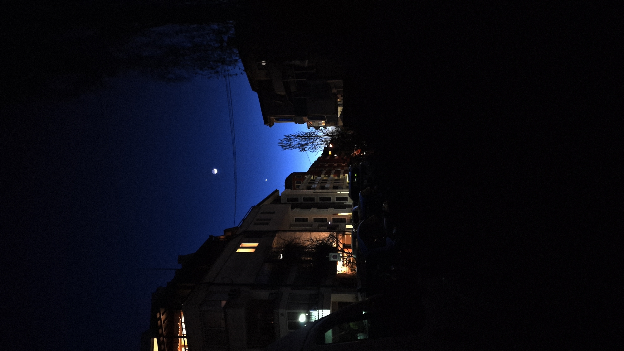 The image depicts a nighttime scene in an urban environment, most likely Sofia, Bulgaria. The camera is tilted, showing buildings rising on the left side. Several buildings with lighted windows are visible, suggesting residential or commercial use. The sky is dark blue, with a crescent moon and a possible star or planet visible. There is a dark, silhouetted area on the right, obscuring detail. A car is partially visible at the bottom. The angle and lighting suggest the photo was taken from street level. There is a tree with bare branches.