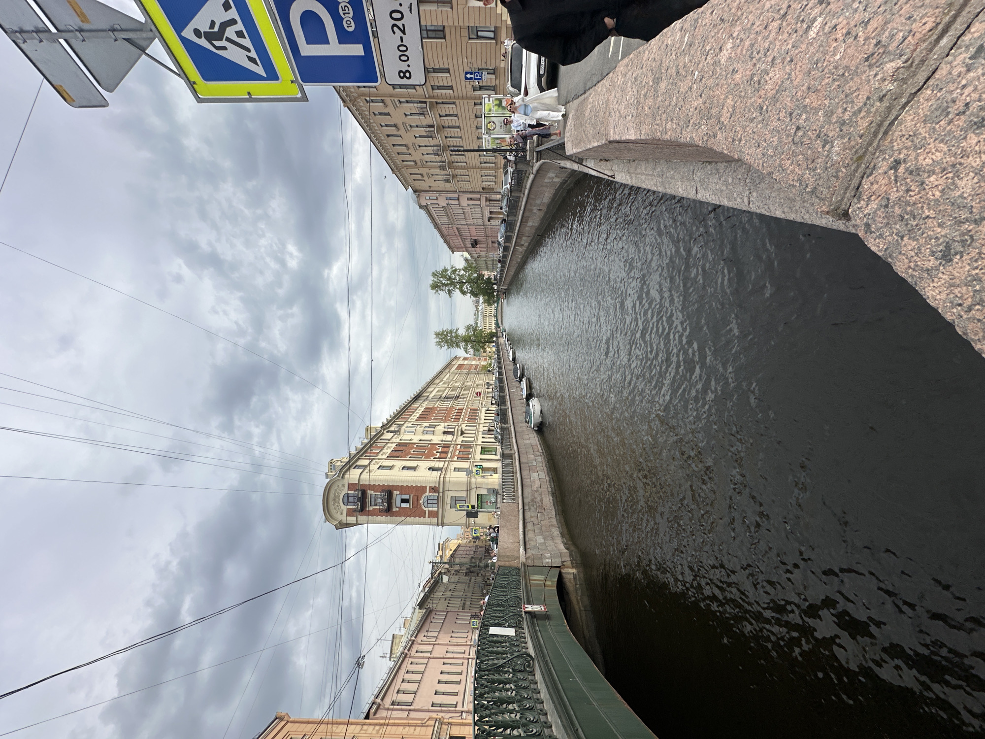 This image depicts a canal in Saint Petersburg, Russia, from an elevated perspective, likely from a bridge. The canal's water is dark, reflecting the overcast sky. Buildings line both sides of the canal, showing classical architecture with multiple stories and varying colors. Pedestrians are visible on the canal's edge, along with parked cars. Signage, including pedestrian crossing and parking signs, are visible on the left side. Power lines stretch across the sky above the buildings. The bridge's edge is visible in the foreground, constructed from granite or similar stone.