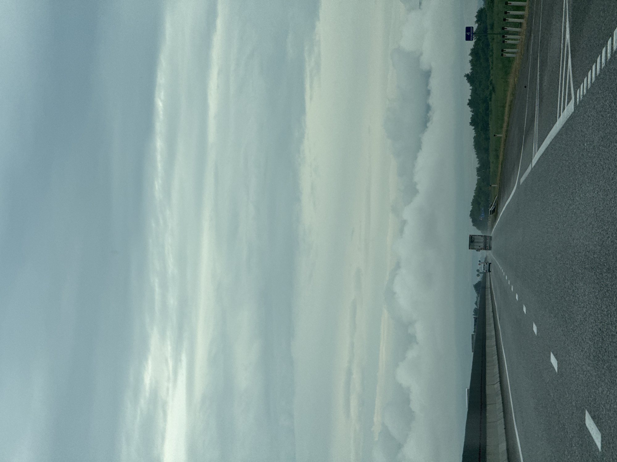 The image depicts a highway scene on an overcast day. A two-lane asphalt road stretches into the distance, marked with white dashed and solid lines. On the left side of the road, a grey concrete barrier separates the highway from what appears to be a grassy shoulder. On the right side, a row of white posts with red reflectors lines the edge of the road, and beyond that, a dense green treeline is visible. In the distance, several vehicles are present on the highway, including a large truck and a car. The sky is filled with thick, grey clouds, creating a diffused light. A blue sign with white text indicating "4 BOGORODSKOE" is visible on the right side of the road. The location is identified as Kochubeyevskoye, Russia.