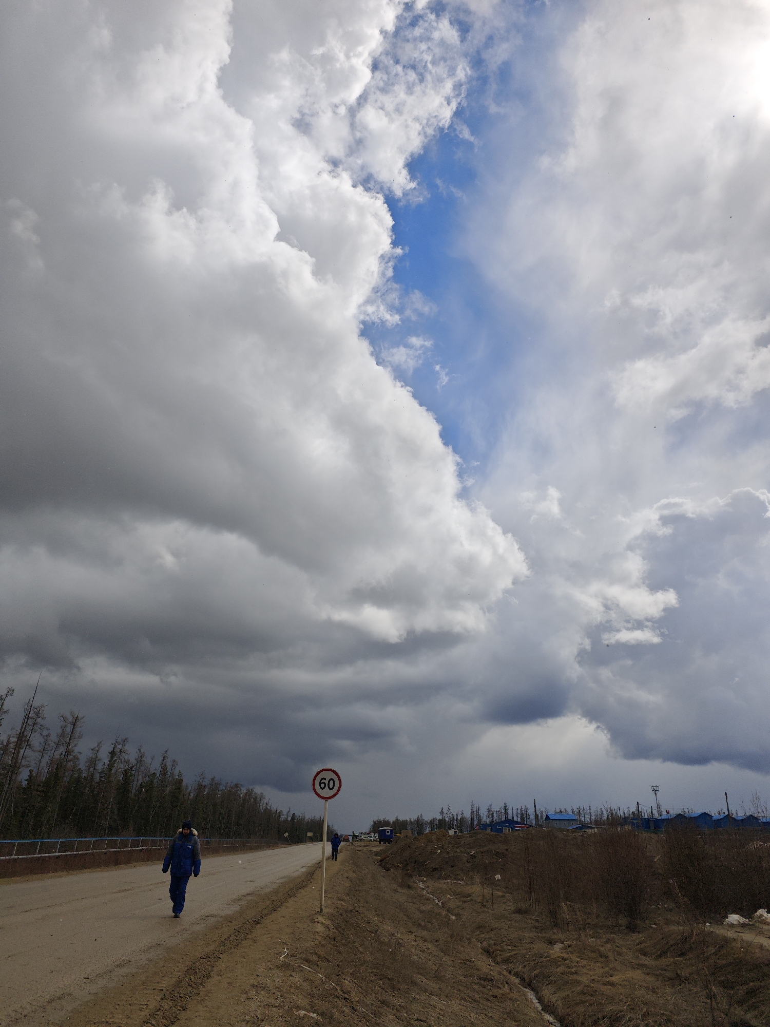 The image depicts a desolate scene in what appears to be a rural area in the city of Unknown, Russia. A man in a blue jacket and dark pants is walking along a muddy road. The road is bordered by a low, rusted metal fence on one side and a barren, brown field on the other. A speed limit sign displaying "60" is visible near the roadside.
In the background, there is a sparse forest of bare trees and a few buildings with blue roofs. The sky is dominated by heavy, gray clouds with patches of blue sky visible. The overall impression is one of bleakness and isolation.
