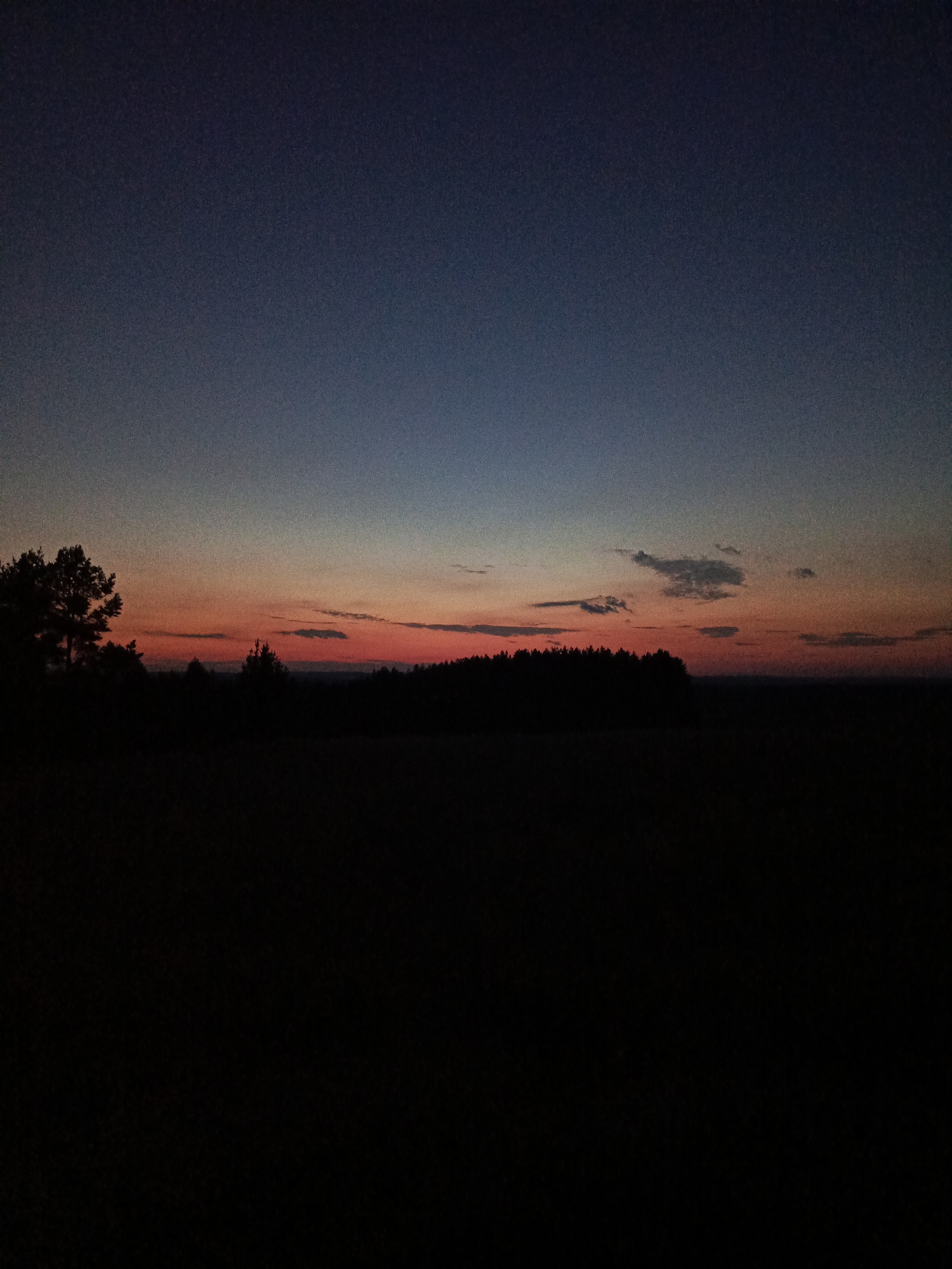 The image depicts a twilight sky with hues of deep blue, fading into orange and red near the horizon. A dark silhouette of a forest lines the horizon, with a single tree visible on the left side. The foreground is a dark, indistinct field. The photograph appears to be taken in an outdoor, rural setting. There are no people or man-made structures visible. The image does not contain any specific visual cues to pinpoint its location within Chumna, Russia.