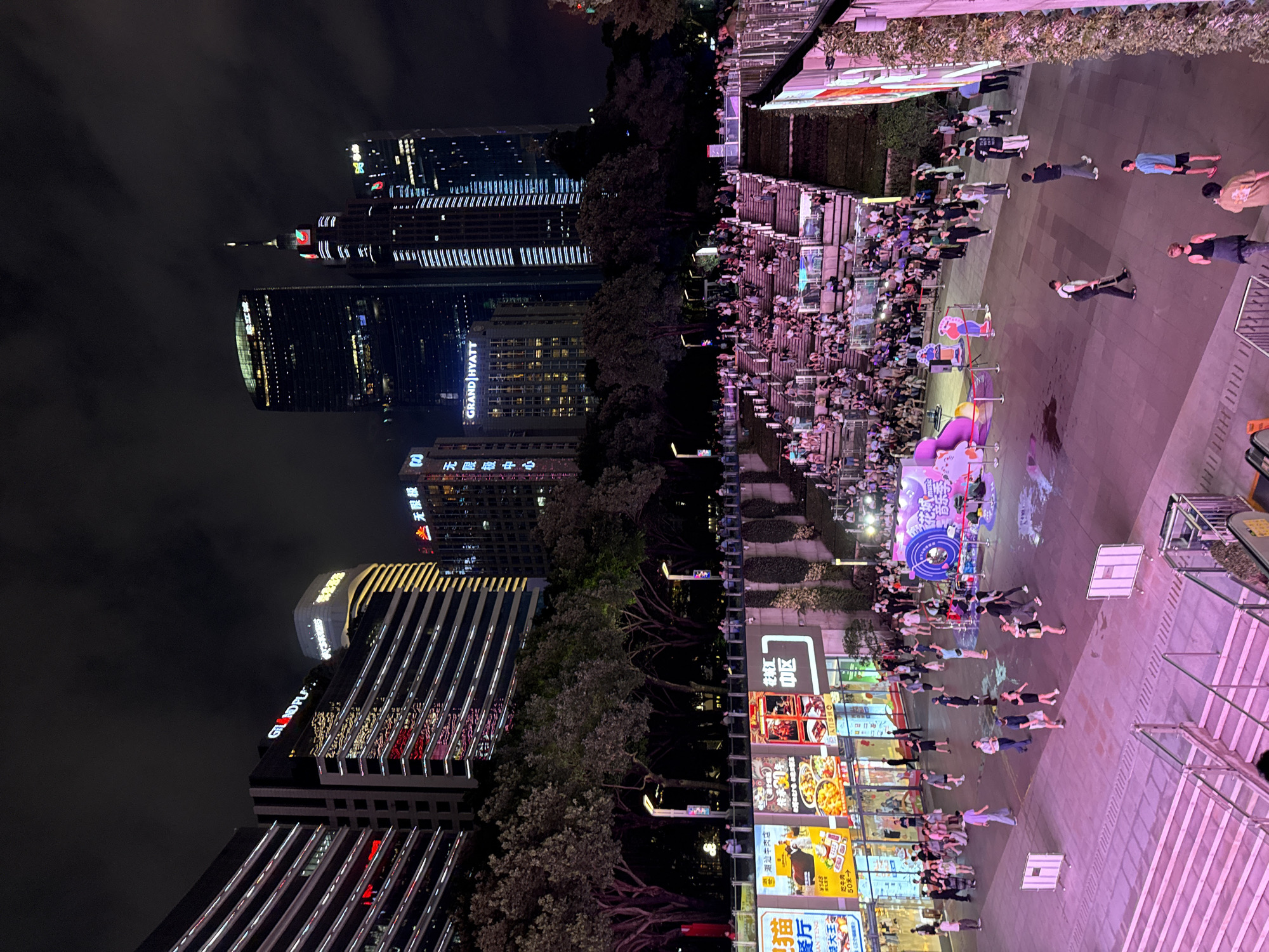 This media file captures a vibrant night scene in Tianhe District, China. The image is divided, showing a juxtaposition of modern urban architecture and a lively outdoor gathering. On the left, towering skyscrapers, some illuminated with the "GRAND HYATT" and "GR NDPL" logos, pierce the dark night sky. Their facades are lined with rows of lights, creating a striking visual contrast.
On the right side of the image, a bustling plaza is filled with people. They are gathered on stepped seating areas, suggesting an event or performance is taking place. Below them, a wide, illuminated walkway is populated by individuals, some standing and others lying down in a synchronized manner, possibly participating in a flash mob or a coordinated activity. Colorful advertisements and signs with Chinese characters, including "花城汇" (Huacheng Hui) and "潮汕牛肉店" (Chaoshan Beef Restaurant), are visible, indicating a commercial area. The lighting suggests it is nighttime, and the atmosphere appears lively and energetic. The overall scene depicts a blend of urban modernity and a dynamic social gathering in a well-lit public space.