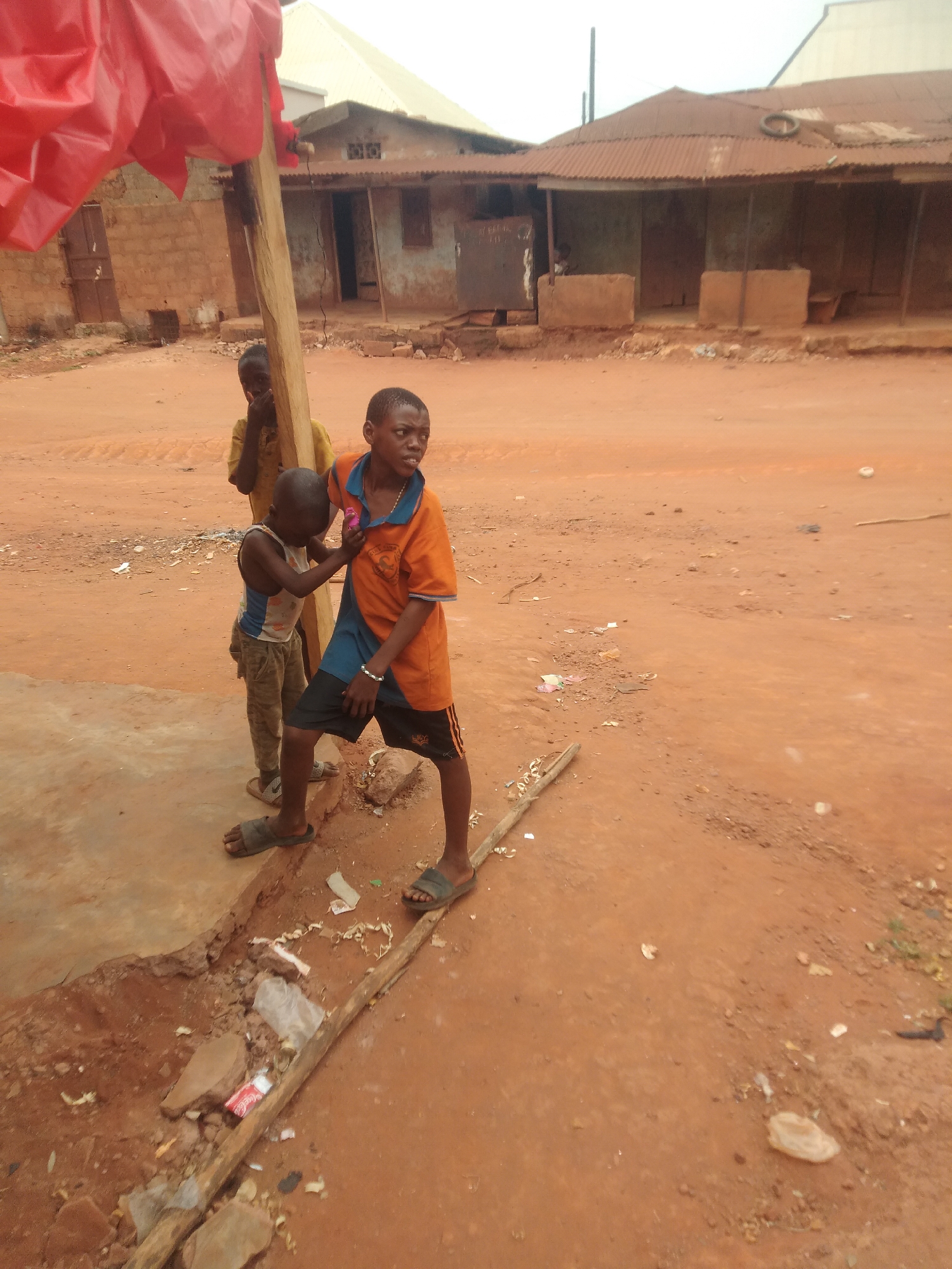 Three boys are on a red dirt road in Ejule, Nigeria. One wears an orange and blue shirt and black shorts, one wears a blue and white tank top and tan pants, and the third is wearing a yellow shirt. All three boys are standing near a wooden post with a red tarp above it. The road is covered in litter. There are buildings with rusted roofs in the background.