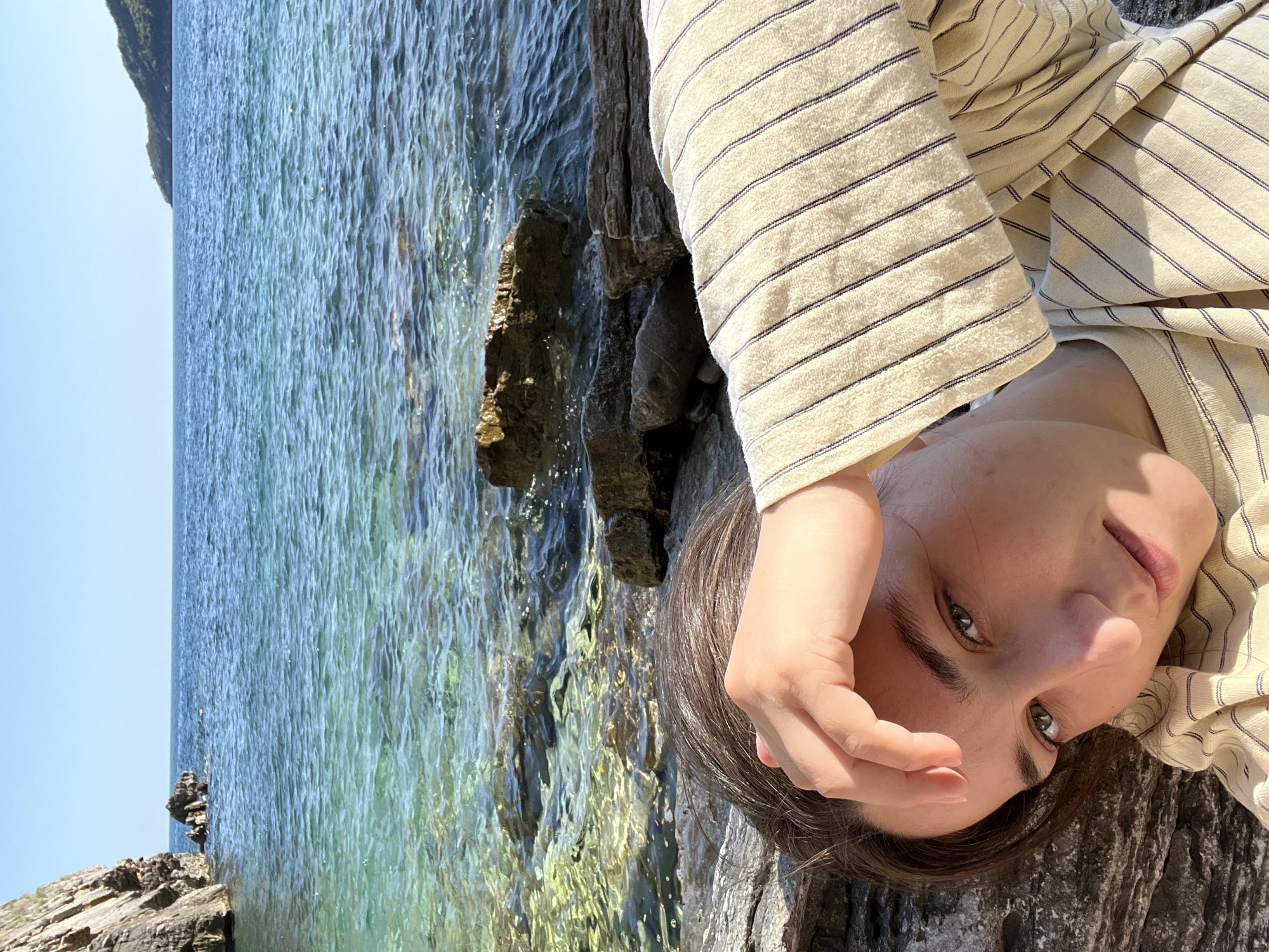 A woman with brown hair and green eyes is lying on a rocky surface near the water. She is wearing a striped shirt. Her right hand is raised near her face. The water is clear and blue, and there are rocks both in the water and along the shoreline. The sky is visible in the upper left corner of the image and appears clear. The location is Budva, Montenegro, as indicated by the rock formations and turquoise sea commonly found there.