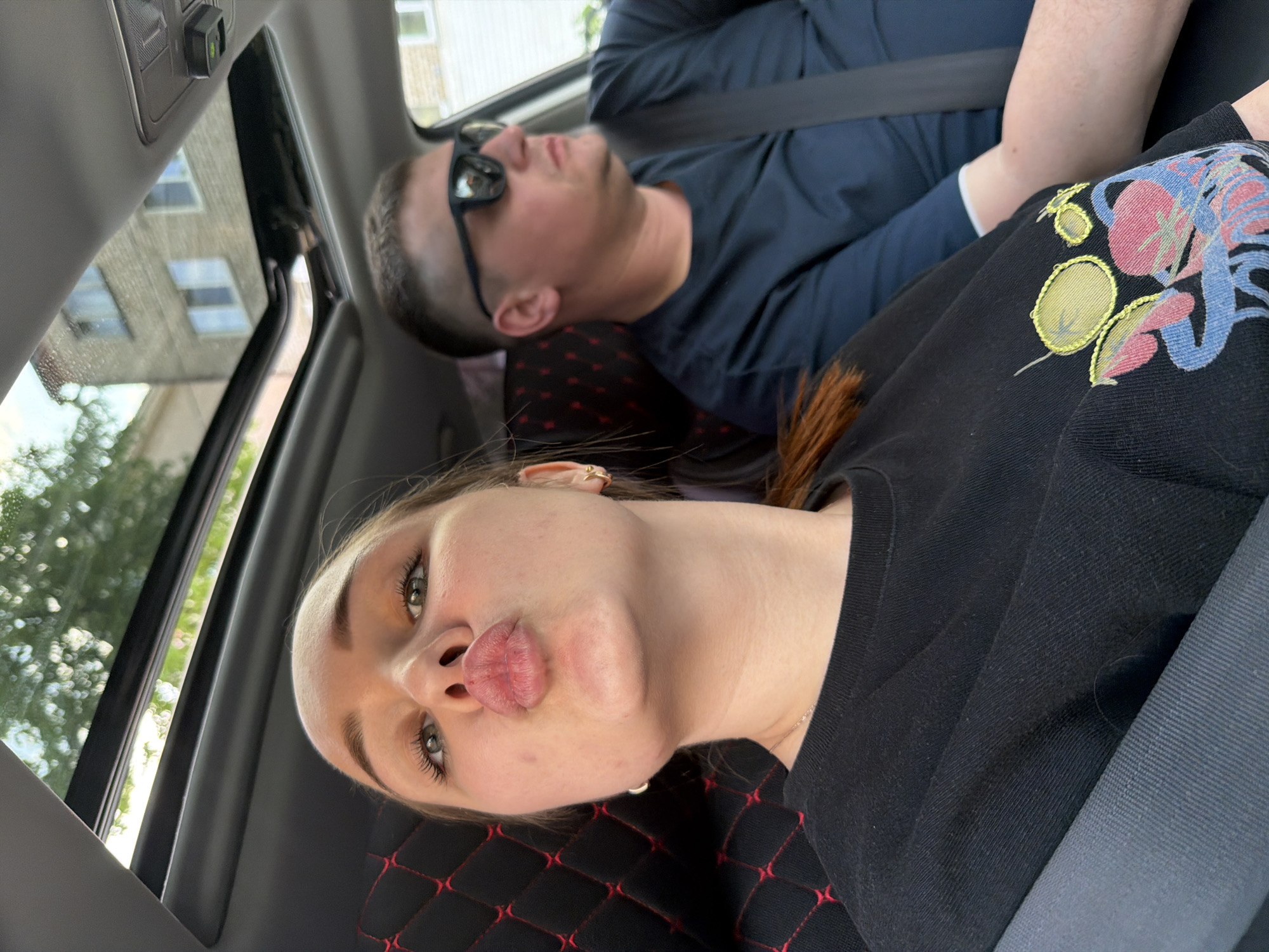 Inside a car, a woman is lying down with a puckered mouth expression towards the camera. She is wearing a black shirt with a colorful design on the shoulder and a seatbelt is fastened across her. Behind her, a man is also lying down with sunglasses on and a blue shirt. The car appears to have a sunroof. Outside the sunroof, a building with windows is visible. The location is not explicitly discernible as Chaikovsky, Russia, only based on the image.
