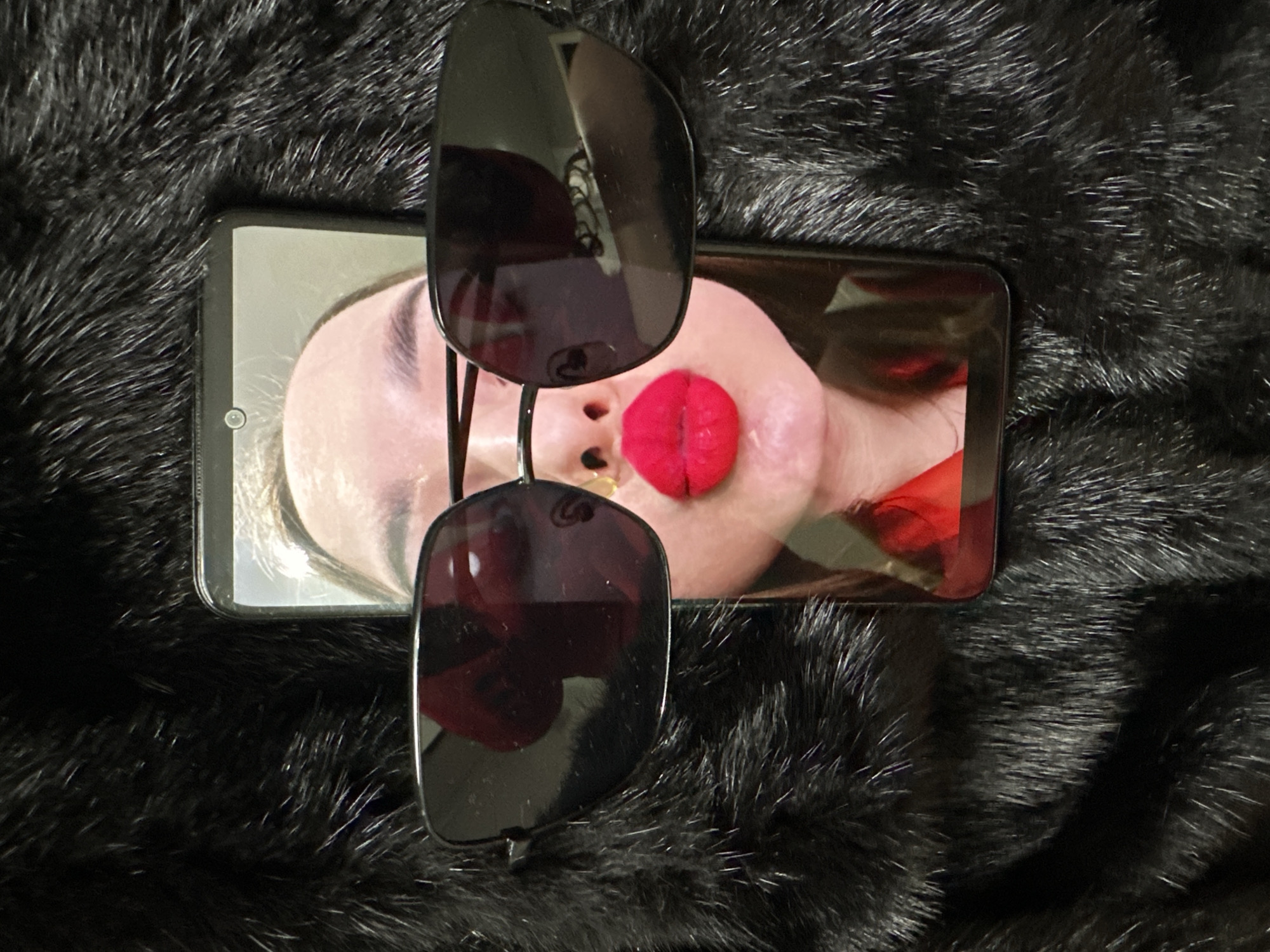 A cellphone rests on a black fur-like surface. The screen displays an image of a person's face with a red object on the nose, partially obscured by a pair of sunglasses. The location in the city of Chaikovsky, Russia is not discernible from the image.