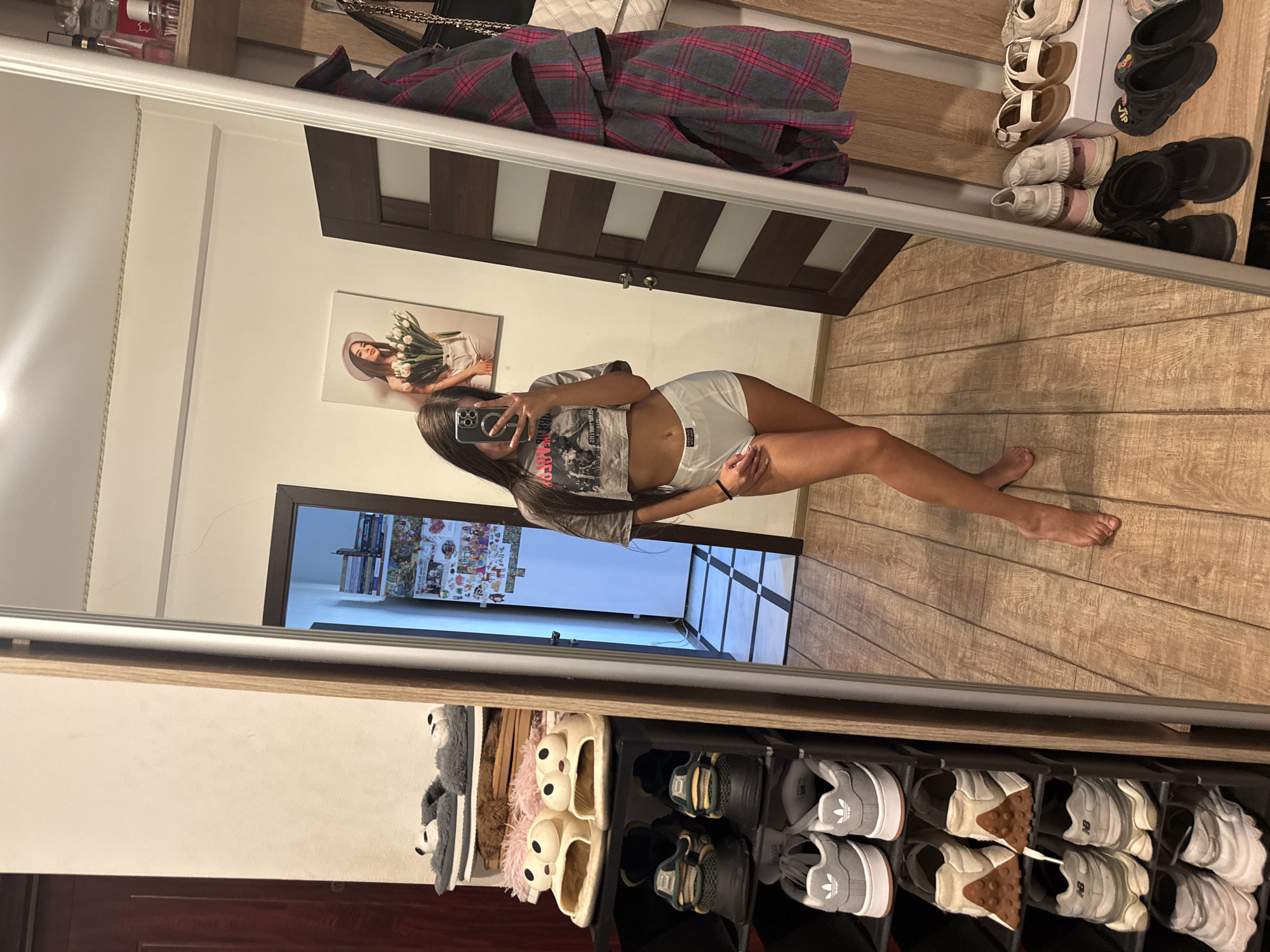 A young woman is taking a selfie in a mirror inside a room. She is wearing a cropped t-shirt and white shorts. She is standing in front of a shoe rack filled with various shoes and slippers. To her right, a plaid jacket is hanging on a hook, and a white purse is also visible. In the background, a door with glass panels is visible, along with a refrigerator covered in stickers and photos. A framed picture of a woman holding flowers hangs on the wall above the refrigerator. The floor is made of wooden planks, and the room appears to be a bedroom or dressing area. The overall scene suggests a casual, domestic setting, possibly in an apartment in Chișinău, Moldova, given the context of the request.