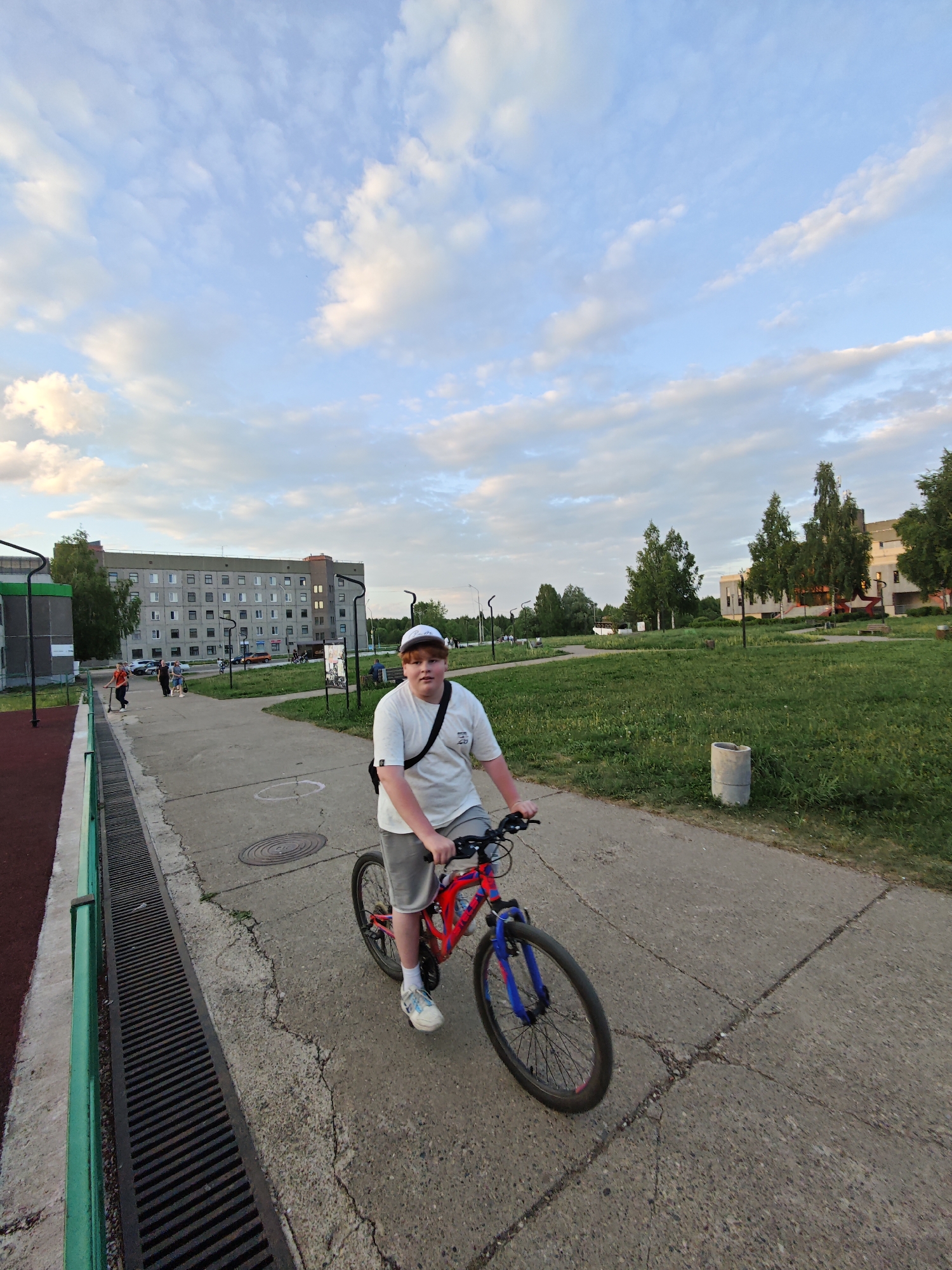 A young male is riding a bicycle on a cracked concrete path in Марковский, Russia. The bike has a red frame and blue front forks. The boy wears a white T-shirt, gray shorts, a white cap, and white tennis shoes. He has a dark messenger bag slung across his chest. To his left is a green and gray structure, and to his right, a grassy area with trees and a concrete cylinder. In the background, a large, gray building is visible, with people walking on a path in front of it. The sky is blue with scattered white clouds.