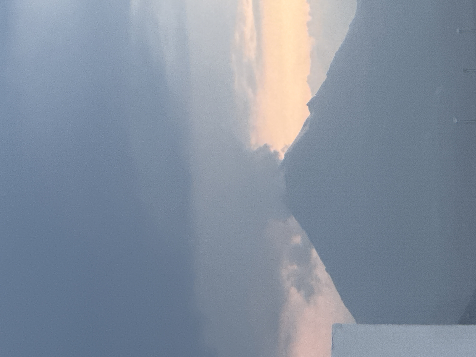 The image displays an outdoor scene, likely a landscape or cityscape, at either sunrise or sunset, characterized by atmospheric haze and dramatic lighting. A large, dark, angular mass, possibly a mountain or a significant man-made structure, dominates the right side of the frame, appearing as a silhouette. Its rugged outline is visible, with faint vertical structures resembling communication towers located on its upper right slope.
The left side of the image features a soft, hazy blue-grey sky that transitions towards the middle. Here, a vibrant source of light, possibly the sun, casts warm yellowish-orange and soft pink hues onto the clouds, creating a striking contrast with the cooler sky. This illumination from behind the dark mass highlights the edges of the clouds.
At the very bottom of the image, a flat, light grey or off-white surface, which appears to be a section of a building's roof or wall, indicates the vantage point is from an elevated position. The overall mood is tranquil and somewhat ethereal, given the soft, diffused light and the atmospheric conditions, suggesting a moment of peace in San Francisco Ocotlán, Mexico. No people, specific activities, or visible text are present.