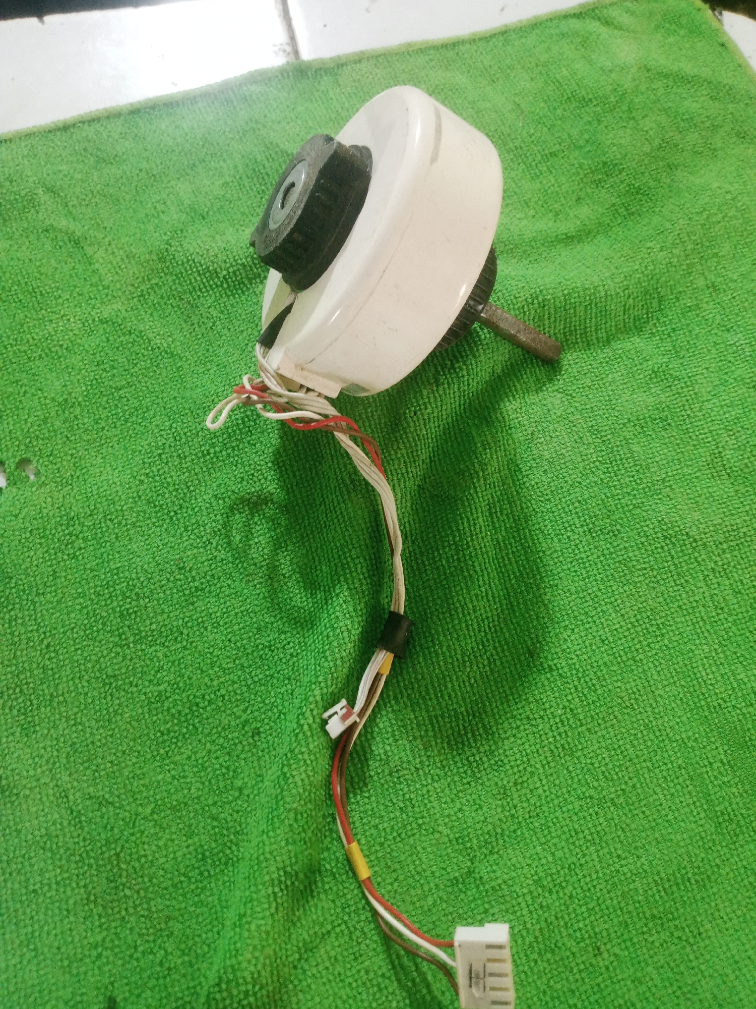 A white motor with a black gear and a metal shaft is lying on a green microfiber towel. Wires with red, white, and brown insulation are attached to the motor, leading to a white electrical connector. The towel is on a white tiled surface. There is no indication of the city of La Tagua, Venezuela, in the image.