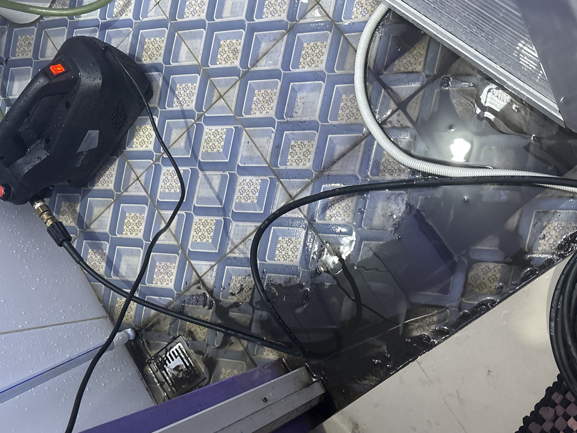 In a bathroom in North Jakarta, Indonesia, a black pressure washer is on a blue and white tiled floor, which is covered in a large amount of dark, murky water. A power cord runs from the pressure washer towards the left side of the image. A hose, possibly connected to the pressure washer, lies in the water. The walls of the bathroom appear to be tiled with white and light blue tiles, and there is a shower drain in the corner, partially submerged in the water. A metallic object, possibly a pipe or a fixture, is visible near the drain. The overall scene suggests a significant leak or overflow in the bathroom.