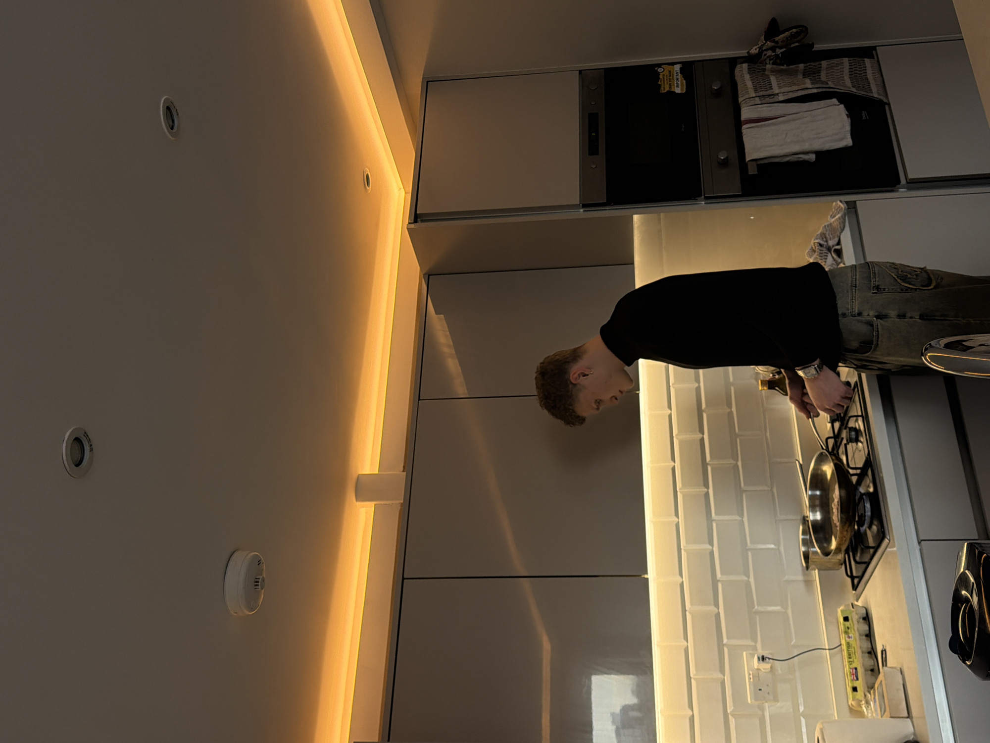 The image displays a male individual cooking in a modern kitchen, identified as being in London, United Kingdom, based on the visible Type G electrical outlet and an egg carton labeled "12 BRITISH."
The male subject, with light brown, curly hair, is seen in profile facing left. He wears a black turtleneck shirt, light-colored jeans, and a metallic watch on his left wrist. His hands are positioned over a frying pan on a black gas hob, indicating he is actively cooking. A smaller silver pot is situated next to the frying pan.
The kitchen features light grey, flat-panel overhead and base cabinets with a white subway tile backsplash. The countertop is a stainless steel surface. Warm amber LED strip lighting illuminates the ceiling perimeter and the area beneath the overhead cabinets. A white smoke detector and a recessed ceiling spotlight are visible overhead. On the backsplash, a white Type G wall socket with a connected white plug is present. A yellow cardboard egg carton, marked "12 BRITISH," rests on the countertop near a roll of paper towels. To the right, an integrated oven/microwave stack displays a yellow "IMPORTANT" sticker, with several kitchen towels hanging from the oven handle. Reflections of an urban structure are visible on the glossy surface of the overhead cabinets. A partially visible black appliance is in the bottom left foreground.