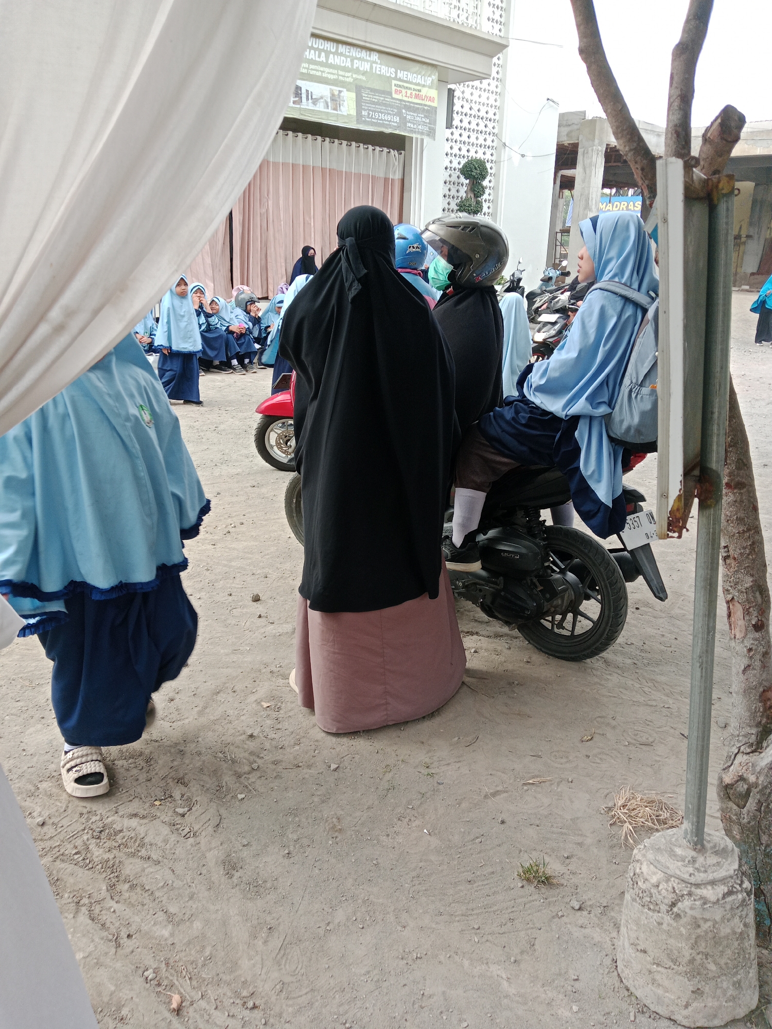 The image depicts an outdoor scene, likely in Kediri City, Indonesia, with a group of people, primarily women and children, wearing Islamic attire. In the foreground, a person in a black hijab and a long pink skirt stands facing away from the camera, with their back towards a parked motorcycle. To the left, another person in a light blue hijab and matching robe is partially visible, also facing away from the camera. Several children in similar light blue attire are seated or standing in the background, some looking towards the left.
The motorcycle, a red scooter, is parked in the dirt, with its rear wheel and license plate (partially visible as "5357 DW") in the frame. The environment is dusty and appears to be an open area, possibly a schoolyard or a public space, with a building and some trees in the background. A white tent or canopy is visible in the far left of the image, partially obscuring the scene. A sign with text in Indonesian is mounted on the building in the background, suggesting it might be an educational institution or community center. A rustic, unpainted metal pole with a concrete base stands to the right of the image, adding to the utilitarian nature of the setting. The overall impression is one of a community gathering or a school event taking place outdoors.