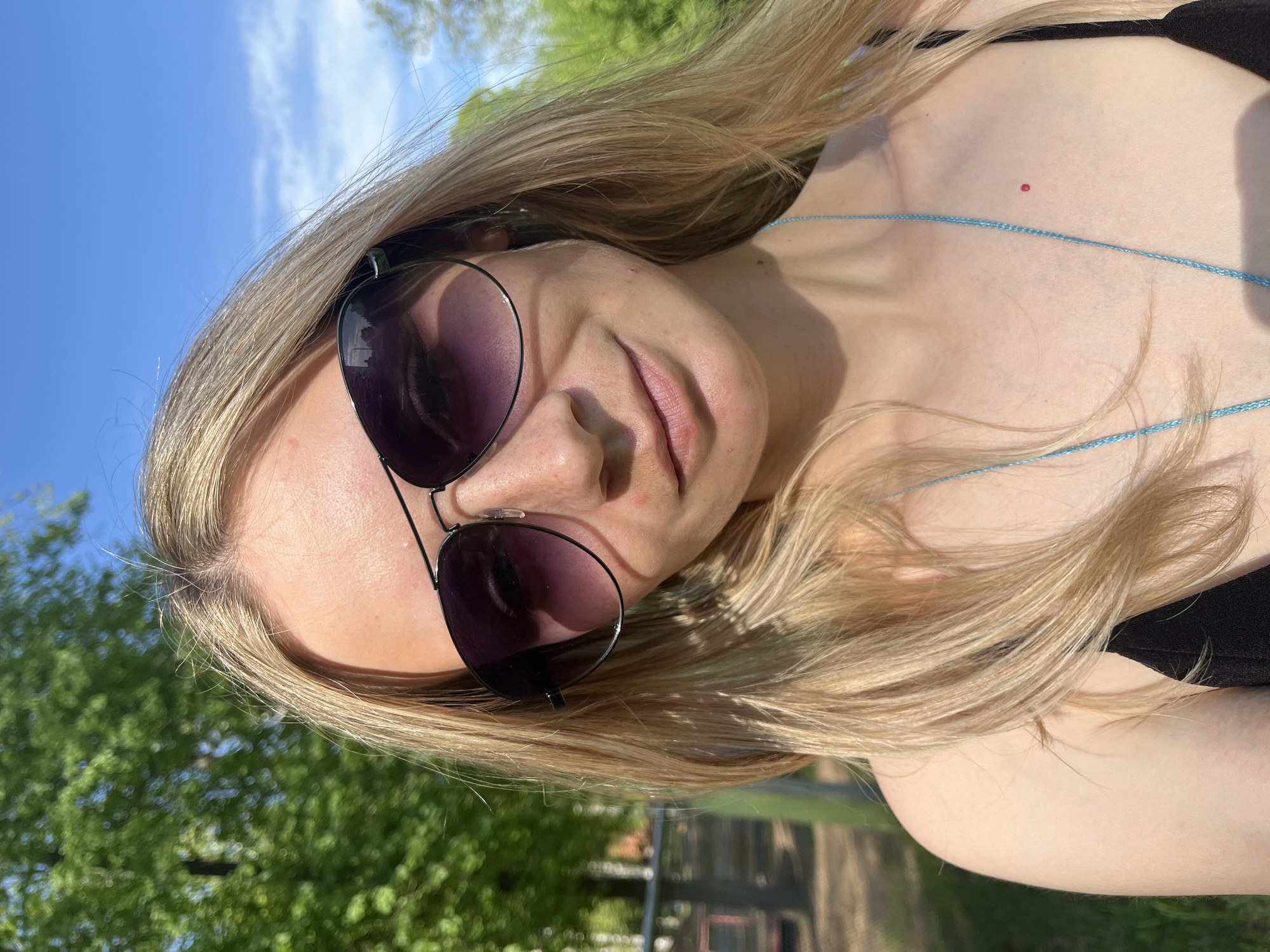 A fair-skinned young woman with blonde hair is wearing sunglasses and a black top. She is outdoors on a sunny day. The location is in the city of Chaikovsky, Russia. Trees and a blue sky are visible in the background. A blue necklace can be seen around her neck. A small red blemish is visible on her upper chest/neck area.