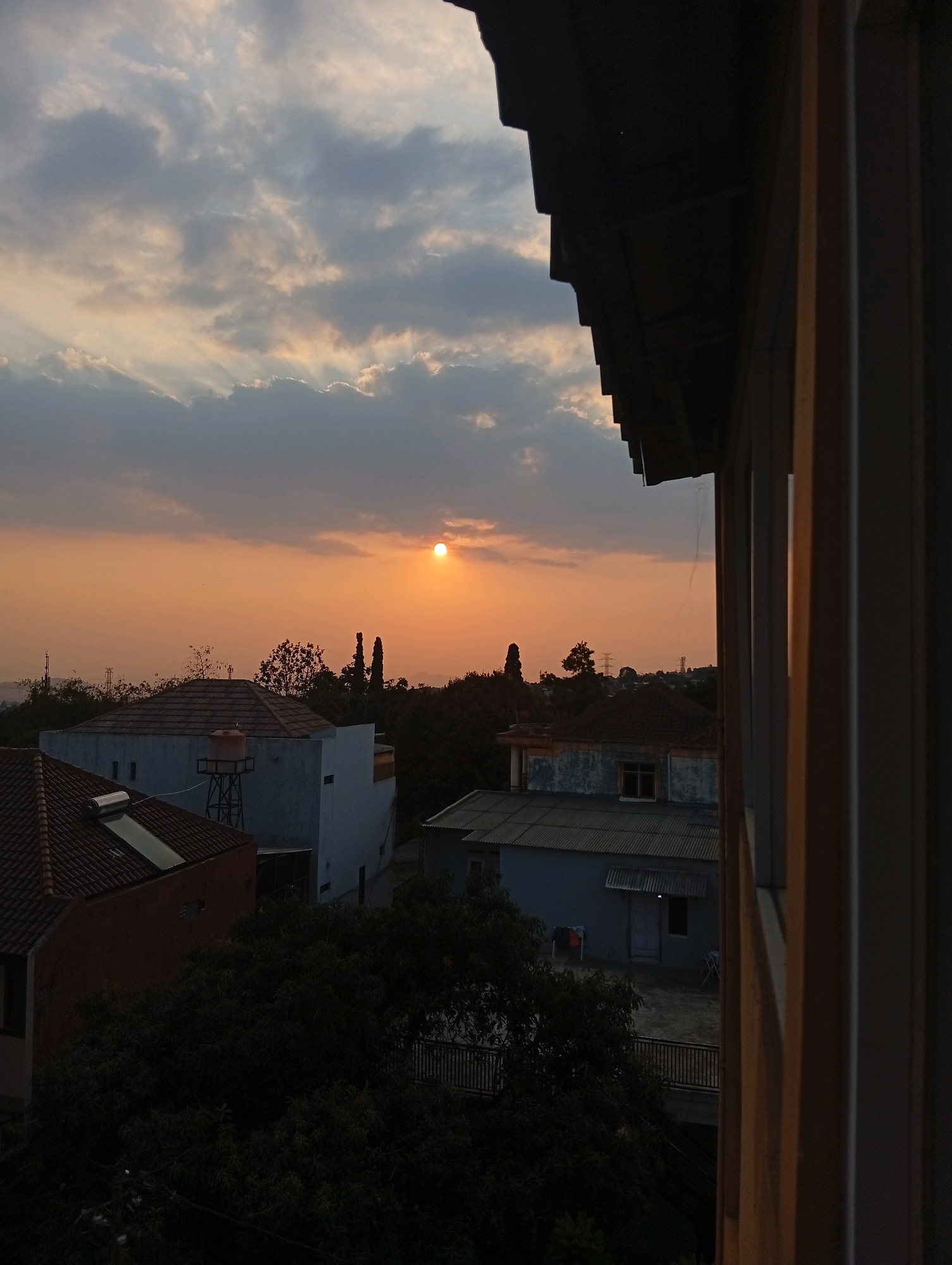 The image depicts a sunset view over a residential area in Bandung City, Indonesia. The sky is a warm orange and yellow, with scattered clouds. The sun is a bright circle near the horizon. In the foreground, the edge of a dark, angled roof structure is visible, casting a shadow. Below the roof, a few buildings with tiled roofs and light-colored walls are scattered among lush green trees. One building has a water tank on its roof. The overall scene is serene and peaceful, typical of a residential neighborhood during sunset.