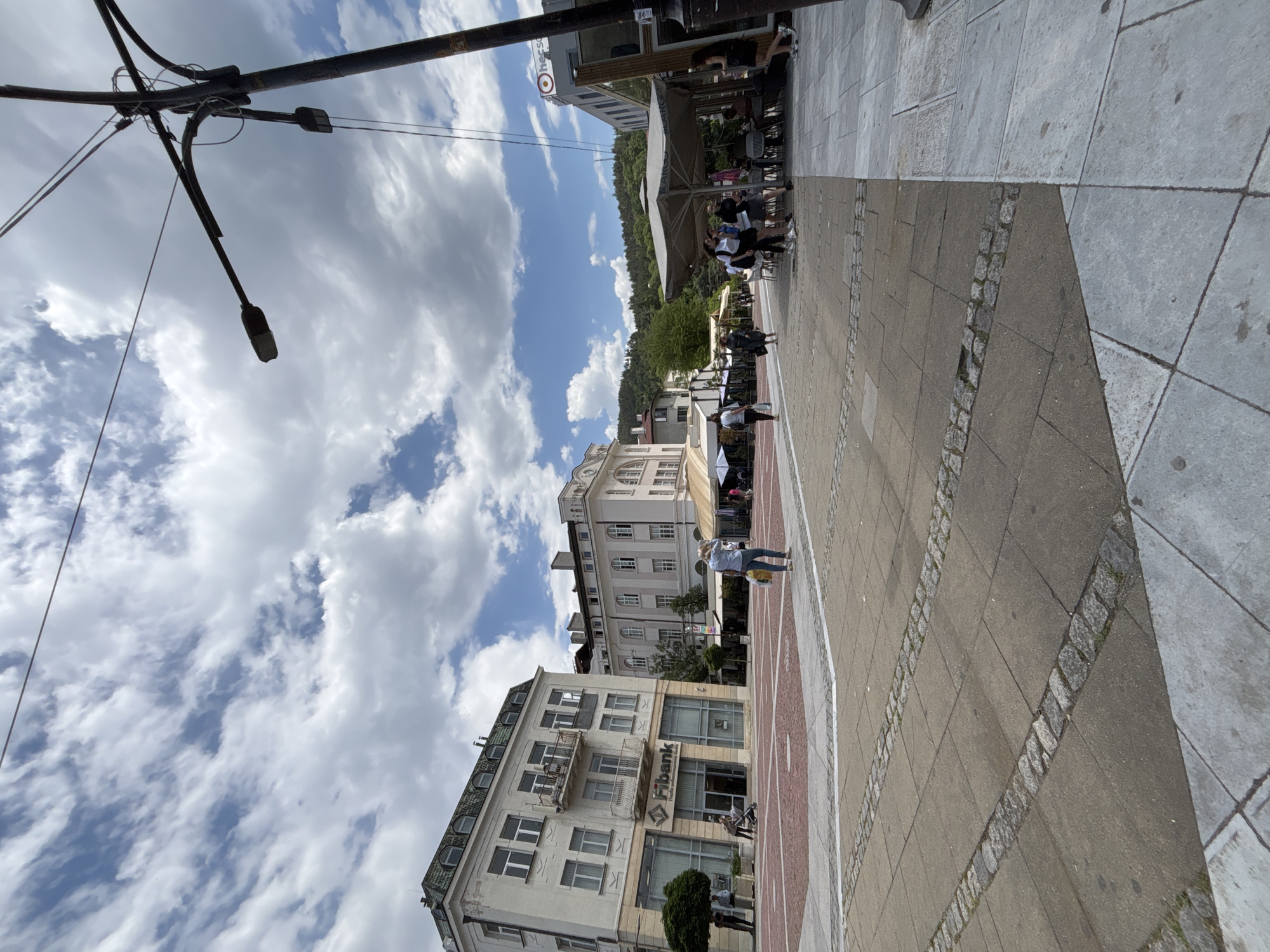 The image depicts a street scene in Blagoevgrad, Bulgaria. The view is oriented vertically, showing the streetscape extending into the distance. Buildings line the left side of the street, including one identifiable as a branch of Fibank. People are present on the street and in outdoor seating areas along the building fronts. The sky is filled with scattered clouds. A street lamp pole is visible in the upper left corner. The street is paved with a combination of stone and concrete slabs.