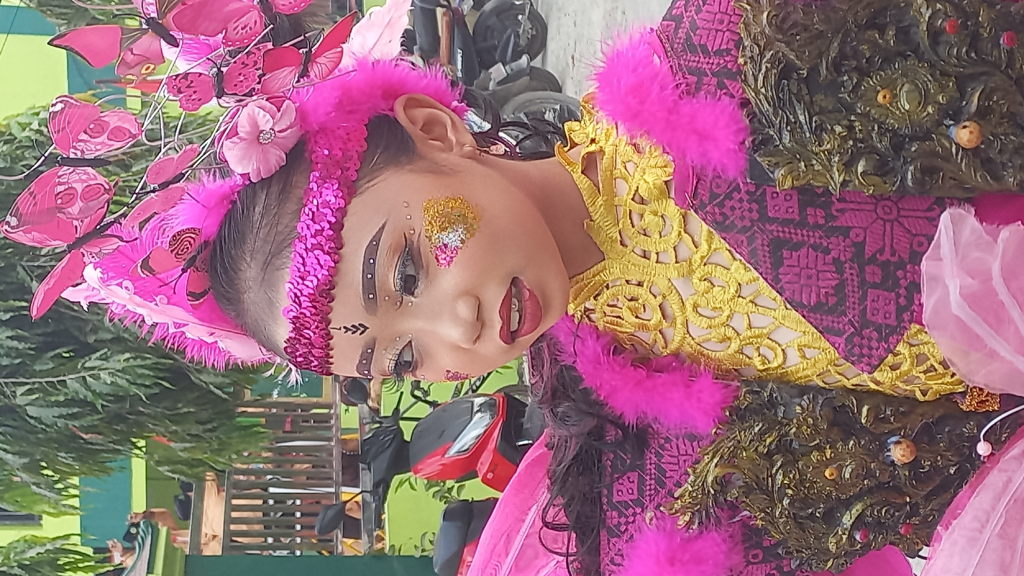 The image shows a close-up of a young girl adorned in a vibrant, elaborate costume, likely for a cultural performance or parade, in what appears to be an outdoor setting. The girl's face is heavily made up with dramatic eye makeup, glitter accents on her cheeks, and dark lipstick. She wears a headdress featuring bright pink feathers and butterfly ornaments. Her costume includes a pink sequined headband, a gold lace collar, a patterned fuchsia garment, and fluffy pink feather trim around her shoulders. Decorative, dark, ornate elements are attached to the front of her outfit. In the background, out of focus, are green buildings, trees, and parked motorcycles. The environment suggests a public space in Kedungmegarih, Indonesia.