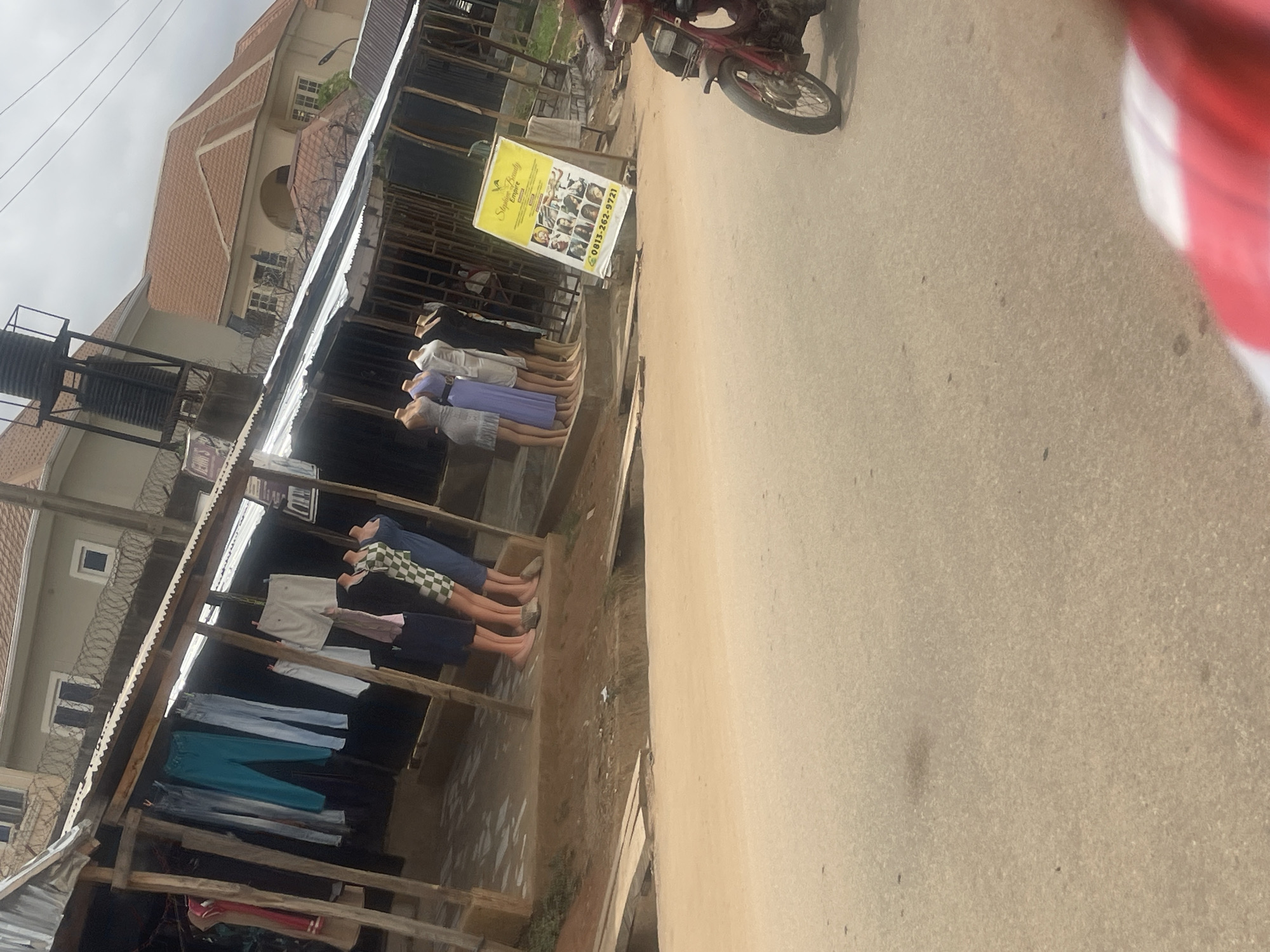 The image depicts a street scene, likely in a market area in Abuja, Nigeria. There is a roadside stall with clothing displayed on mannequins. Several mannequins are seen, with clothes on display, and trousers hanging along the stall. A sign with "Stephen Family" written on it and a phone number is visible in the upper right quadrant. A motorcycle passes on a paved road. A building is visible in the background.