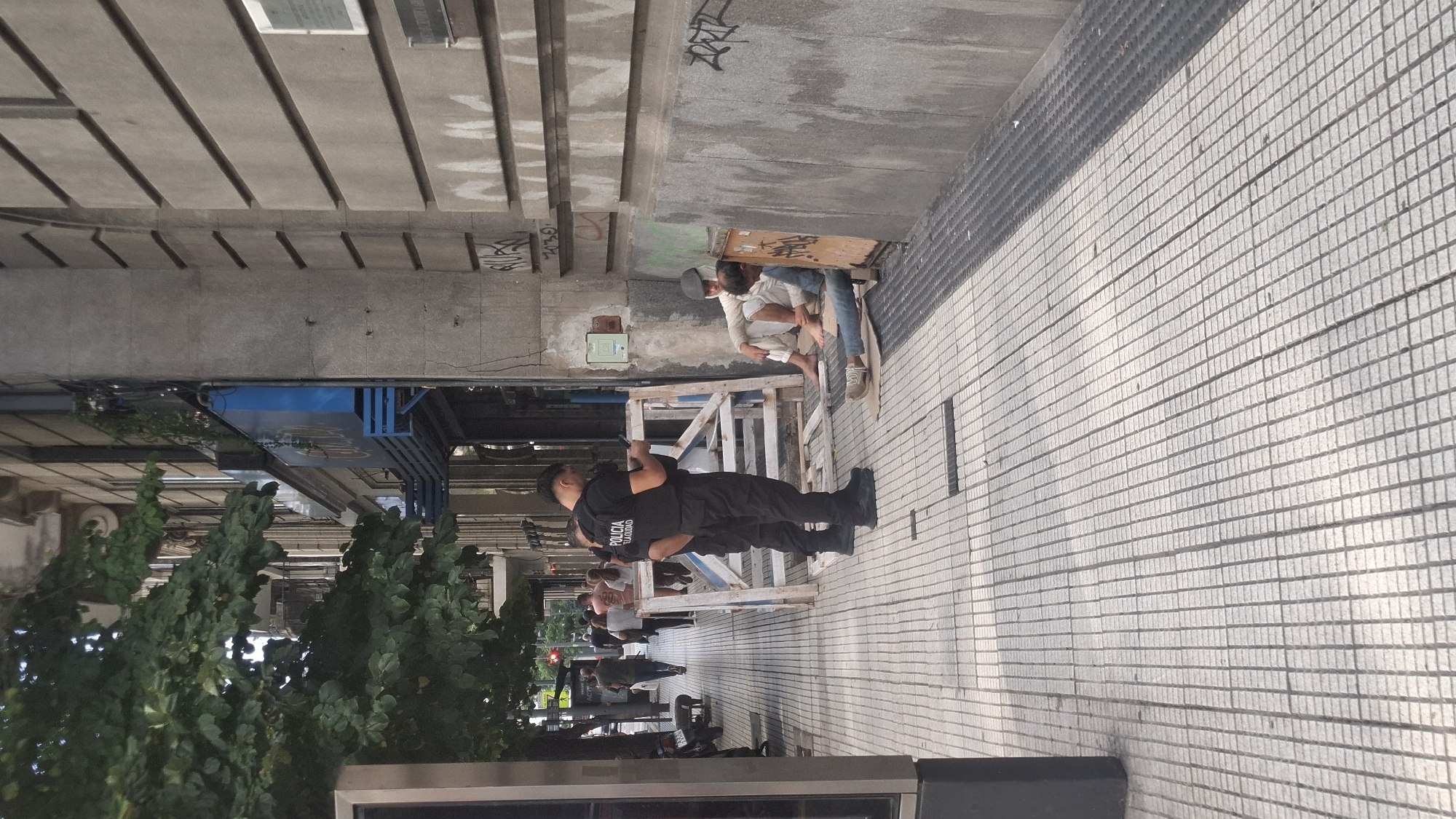 The image depicts a scene on a sidewalk in Buenos Aires, Argentina. Two individuals are seated on the ground with a board above them. A police officer, wearing a uniform that reads "POLICIA CIUDAD", is standing near them, seemingly observing. Construction barriers are visible between the officer and the seated individuals. Other pedestrians are walking by in the background, including the silhouette of another police officer. The environment consists of a sidewalk paved with small, light-colored tiles and a building facade with concrete elements and graffiti. Trees are visible to the left of the frame.