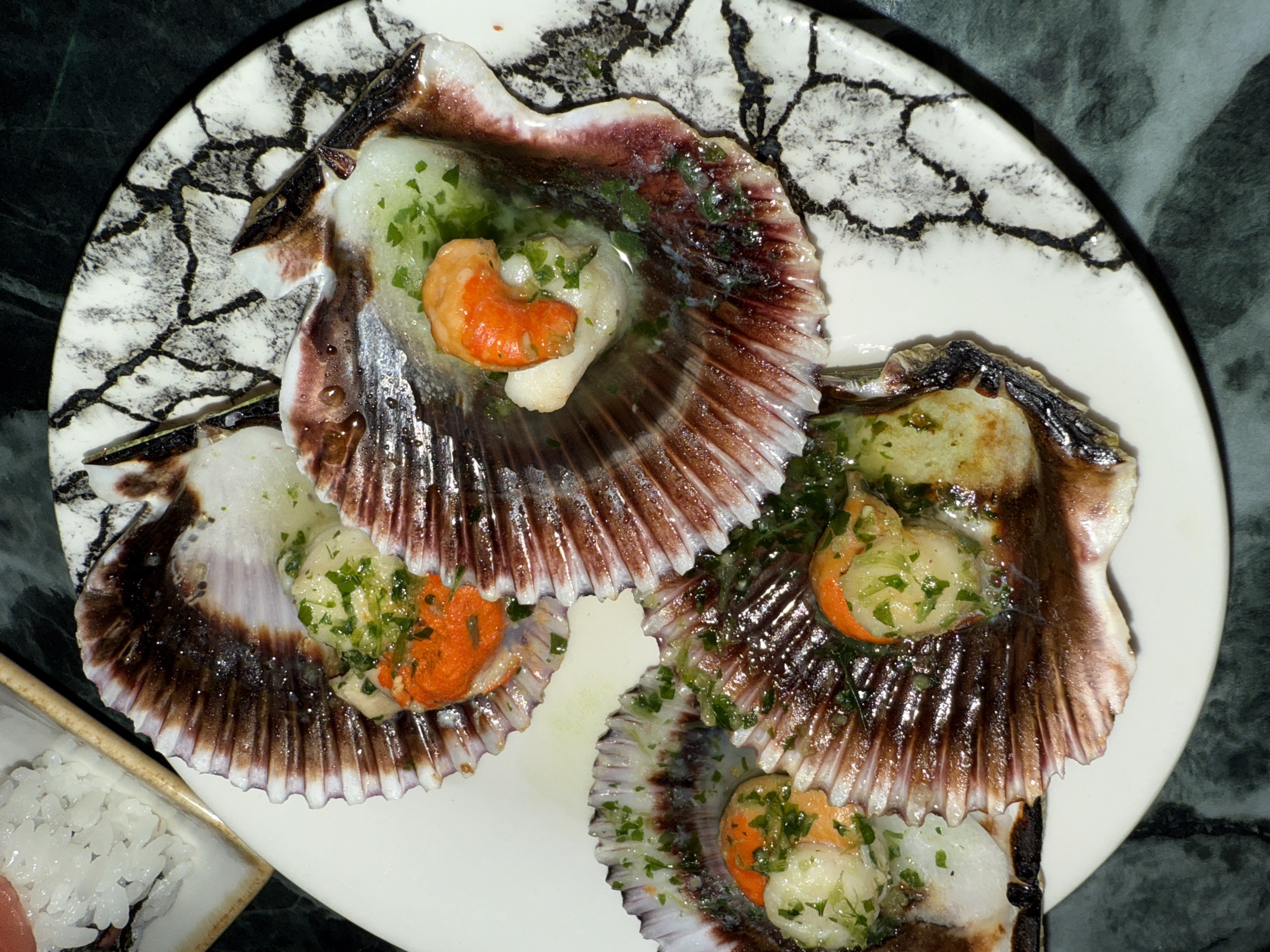 The image depicts a plate of scallops served in their shells, likely in a restaurant setting. The scallops are garnished with herbs and appear to be cooked with some kind of sauce. The plate itself is white with a black crackled pattern. A small dish containing sushi is also partially visible on the lower left side of the frame. The setting appears to be indoors, on a dark green or black marble surface. Given the subject matter and quality of the image, it's plausible that this photo was taken at a seafood restaurant in a city known for its culinary scene, potentially Barcelona, Spain.