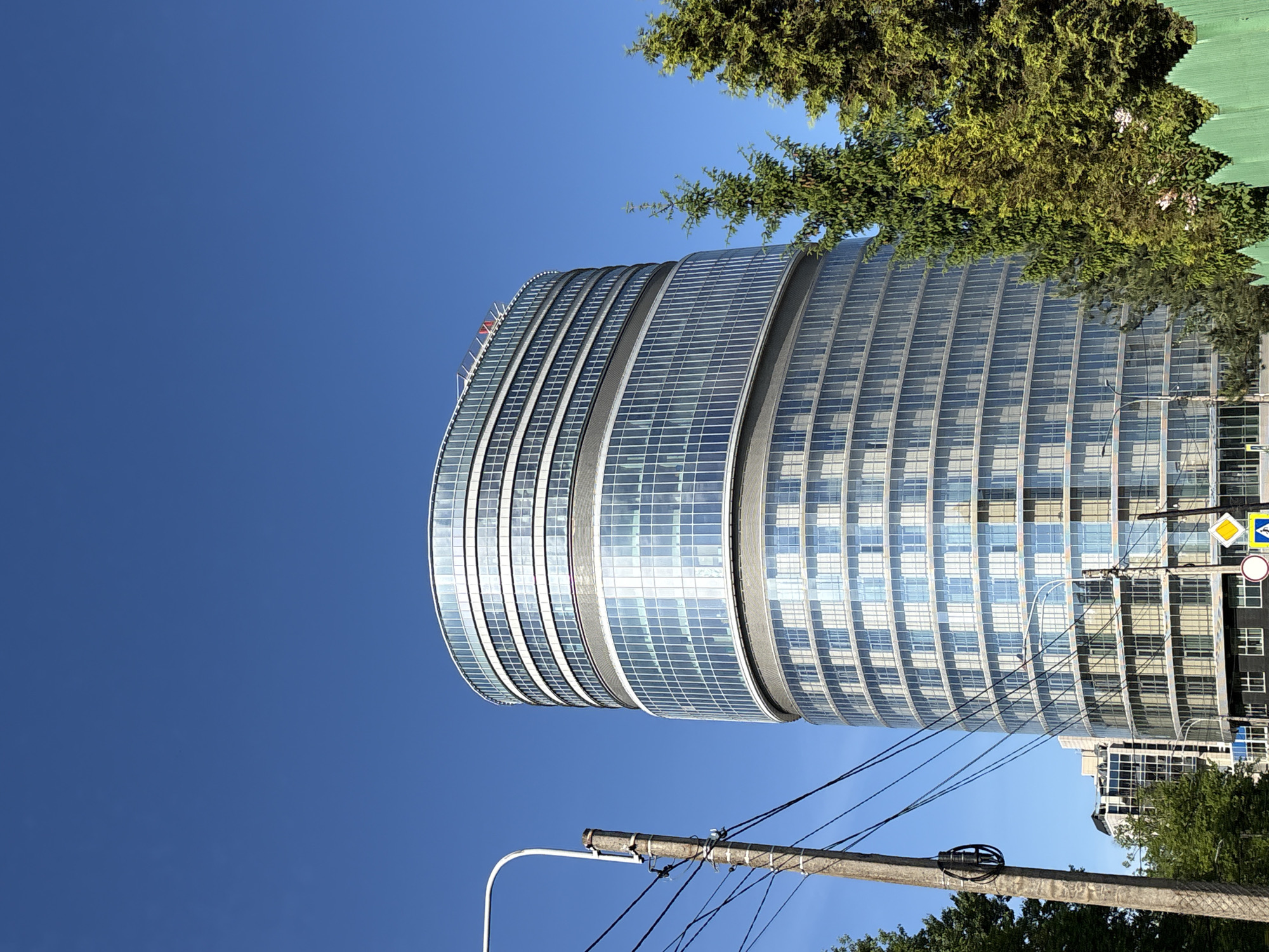The image, viewed upright, showcases a modern urban landscape under a clear blue sky on a sunny day. The dominant feature is a large, contemporary building with a distinctive curved, cylindrical shape. Its facade is composed entirely of reflective glass panels arranged in numerous horizontal bands, creating a sleek, shimmering surface. On the top edge of this building, a red logo or text in Cyrillic letters, appearing to be "ВТБ" (VTB), is visible.
To the upper right of the building, the lush, dark green foliage of coniferous trees extends into the frame. In the foreground, spanning the lower section of the image, is a wooden utility pole laden with numerous black electrical wires that crisscross the view. A white street light fixture is also attached to this pole.
On the right side of the image, near the building and utility pole, several traffic signs are visible: a yellow diamond sign indicating a main road, a blue square sign with a white arrow, and a red circular sign with a diagonal line, typically meaning "no stopping." Partially visible behind the main building is another, less distinct building with dark rectangular windows.
The sky is a uniform, brilliant blue, indicating bright, clear weather conditions. The scene is static, devoid of people or specific activities, capturing a typical street-level view of a modern city area during daylight hours.
