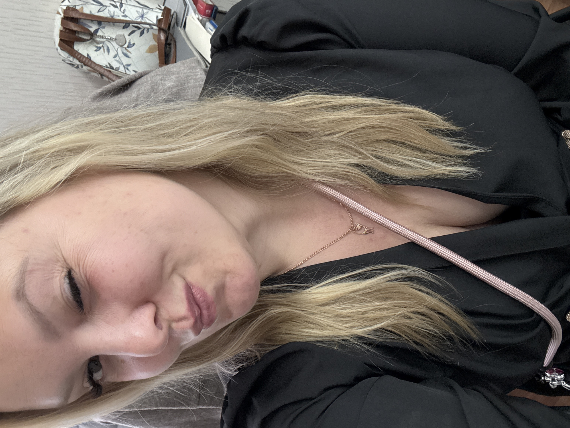 A close-up photograph depicts a fair-skinned woman with blonde hair, wearing a black garment. A necklace is visible on her chest. In the upper left corner, a handbag with a floral pattern is partially visible. The location in the city of Noviy, Russia cannot be confirmed from this image.