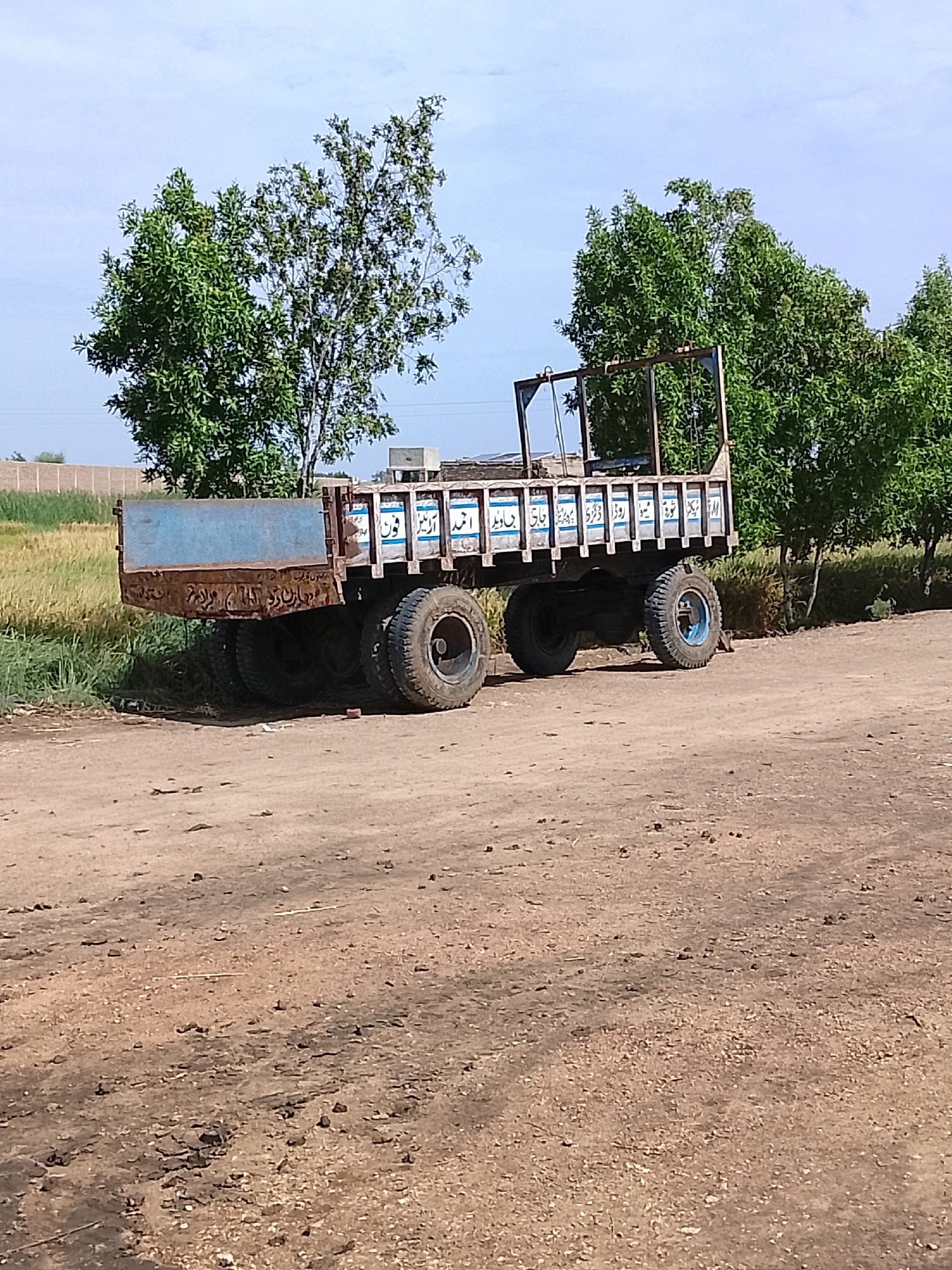 An old, rustic trailer truck sits parked on a dirt road in Pataar Goth, Pakistan. The truck's blue bed is faded and worn, with visible rust along the edges. Arabic script is inscribed on the side of the trailer's wooden planks. The truck is supported by multiple large tires. In the background, lush green trees and a field of wheat are visible under a cloudy sky. The overall scene depicts a rural setting with a focus on the weathered vehicle.