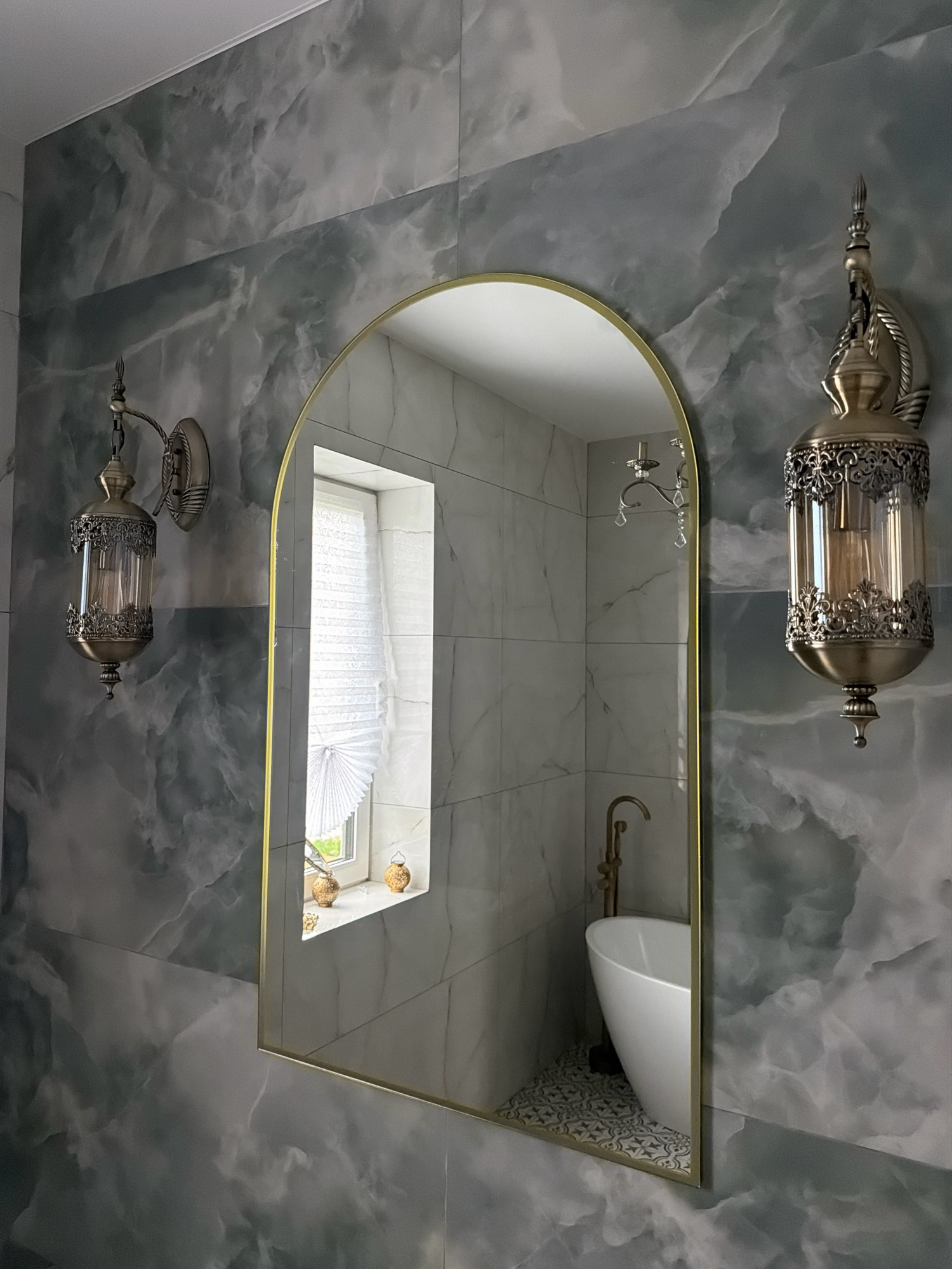 The image depicts a bathroom with dark gray marble-patterned tiles on the walls. A large, arched mirror with a golden frame hangs centrally, reflecting a white bathtub with a gold faucet and a window with a white pleated blind. Two ornate, vintage-style wall sconces with glass shades and metal detailing flank the mirror. One sconce is visible on the left, and its reflection is visible on the right side of the mirror. The floor in the reflected bathtub area has a decorative tile pattern. The scene suggests a luxurious and antique-inspired bathroom design. There are no people present in the image. The location is identified as a bathroom in the city of Orsha, Belarus.