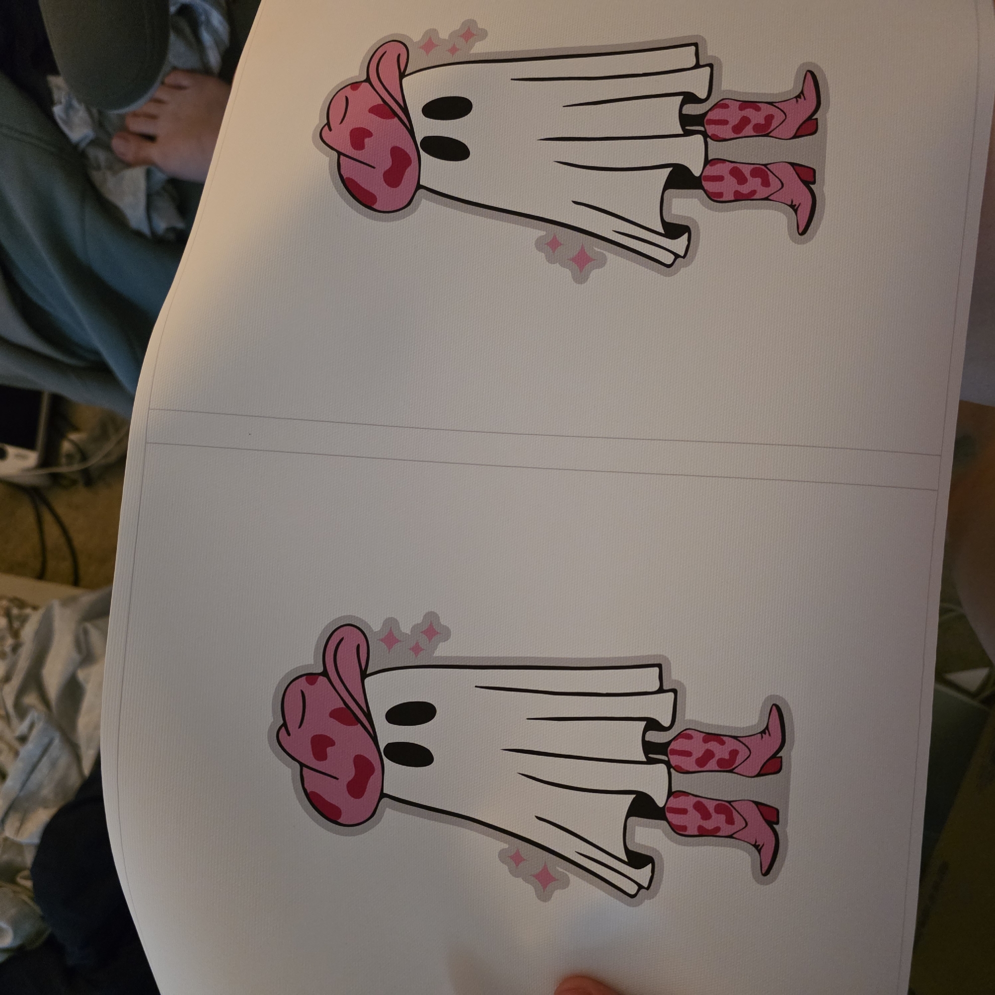 The image displays two identical cartoon illustrations of a ghost wearing a pink cowboy hat and pink cowboy boots. The ghost is white with black oval eyes and a flowing sheet-like body. Pink starbursts are scattered around each ghost, indicating a sparkling effect. The illustrations are printed on a large piece of white paper, which appears to be a poster or print. The paper is partially unfurled and lies on a surface, possibly a floor or table, with some clothing and a bare foot visible in the background, suggesting a casual indoor setting. The scene is not in a specific location in Portland, Oregon, as no identifying landmarks or contextual clues are present. The lighting is diffuse, and the focus is on the printed illustrations.