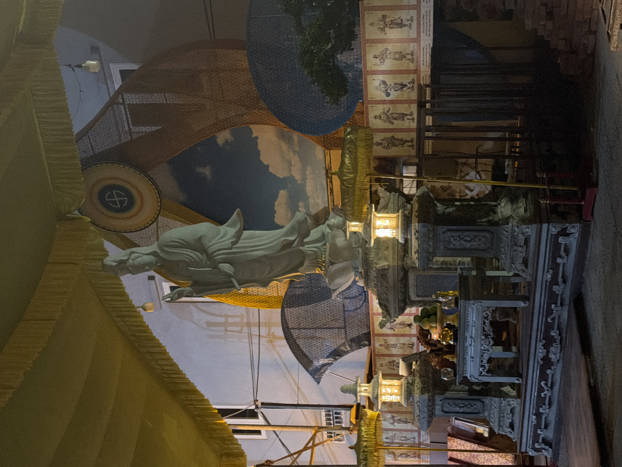 The image depicts a nighttime scene at a religious or cultural site, likely in Đà Nẵng, Vietnam. A large, pale statue of a robed figure, possibly a deity or historical personage, stands prominently in the center, slightly to the right. The statue is elevated on a tiered stone pedestal.
In front of the pedestal, there is a low, ornate table with a bowl of fruit and other small offerings, suggesting a place of worship or remembrance. Flanking the pedestal are two elaborately designed lanterns, one on each side, casting a warm yellow glow. The area is decorated with various cultural elements, including intricate patterns on screens or backdrops, and a large circular emblem with a symbol in the center, positioned behind the statue. Below this, a mural-like scene of clouds is visible.
To the left of the statue, a golden umbrella with tassels is visible, and to the right, a dark metal fence or railing encloses a shadowed area. Along the base of the main structure, there are a series of framed images or paintings depicting figures in traditional attire. The overall lighting suggests it is after dark, with artificial lights illuminating the central display. The background shows architectural elements and electrical wires, indicative of an urban or semi-urban setting. The ground in the foreground is dark and appears to be paved or concrete. The presence of multiple decorative elements and the statue strongly suggests a temple, shrine, or cultural performance venue.