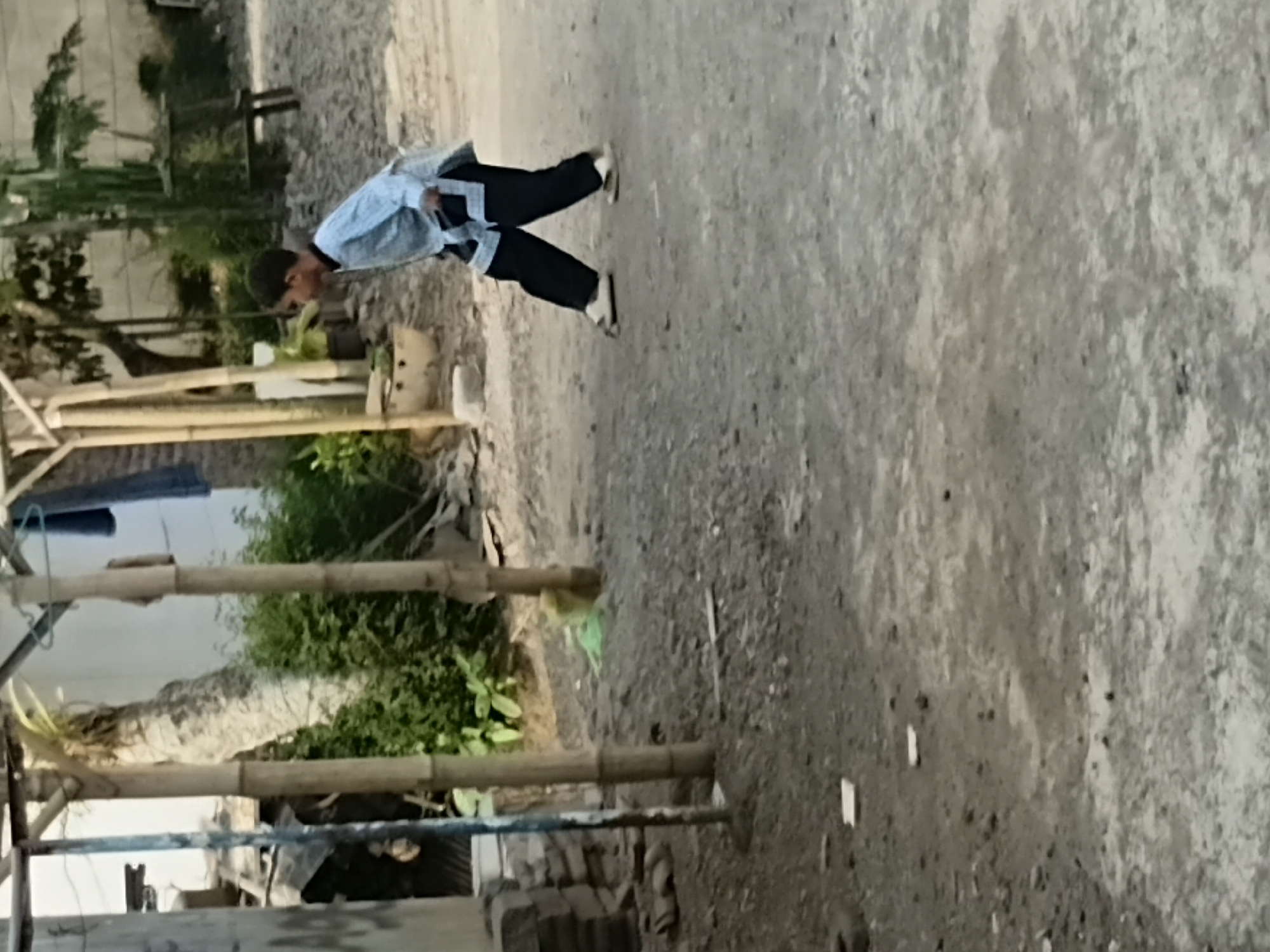 In Tanggul Wetan, Indonesia, a young boy dressed in a light blue shirt with a dark collar and dark pants with white accents walks across a dusty, uneven ground. The boy is in motion, with his right leg forward and his left leg trailing. He is wearing light-colored sandals. Behind him, bamboo poles support a makeshift structure, and lush green foliage is visible. The background includes a white building and more greenery. The overall scene suggests an outdoor setting, possibly a residential area or a community space.