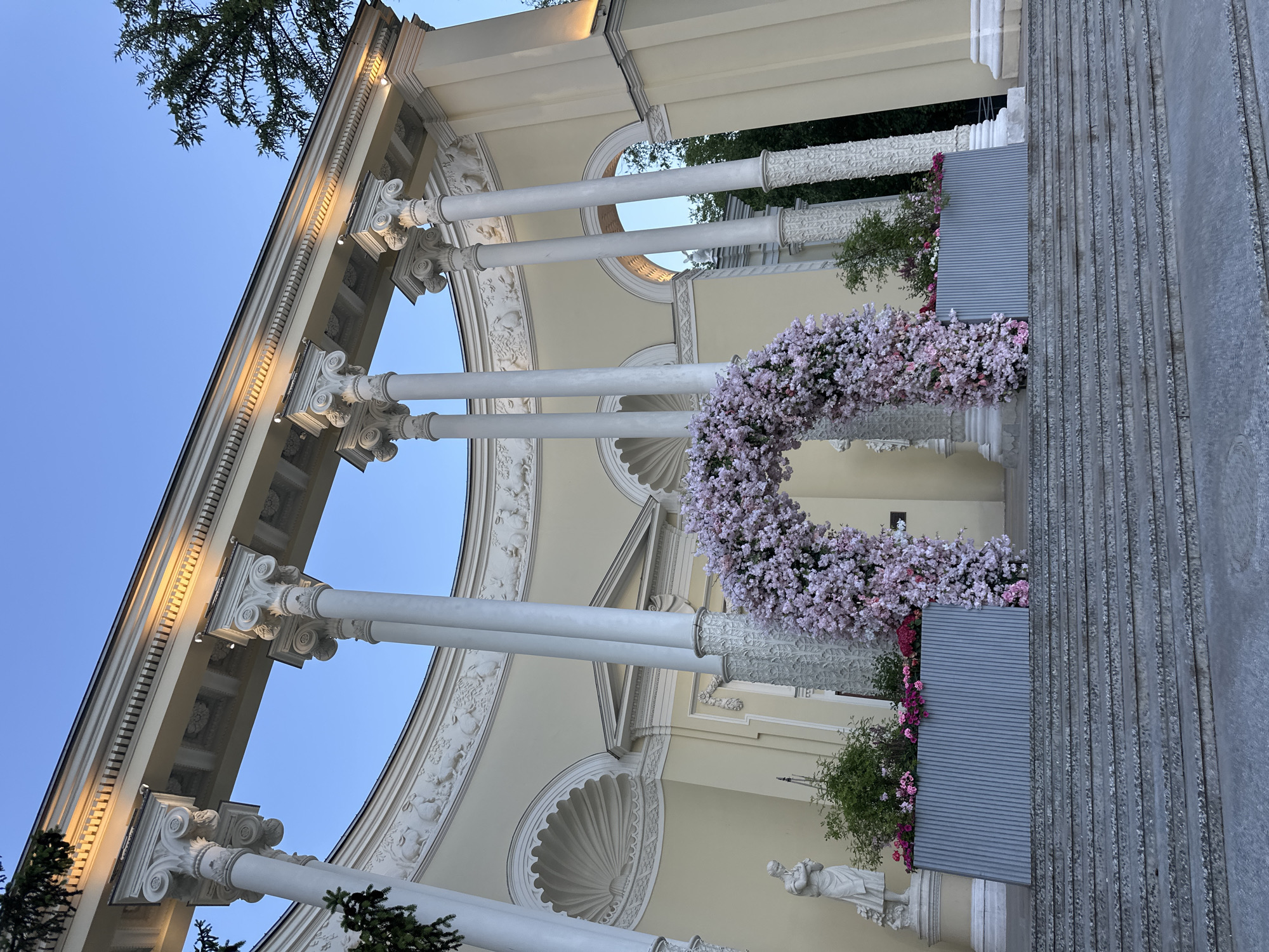 The image displays an ornate classical-style pavilion or colonnade, consistent with structures found in historic parks or estates, such as those in Moscow, Russia. The architecture features numerous tall, white fluted columns, topped with elaborate Corinthian or Composite capitals, supporting a cream-colored entablature. This entablature is richly decorated with bas-relief carvings depicting figures and floral motifs. Rounded archways with decorative shell-like elements are visible between the columns.
A prominent focal point is a large, heart-shaped (or U-shaped) floral arch, densely covered in small pink and white flowers, positioned within the structure. Additional planters filled with green foliage and smaller pink/red blooms are arranged around the base of the columns. The underside of the entablature is illuminated by artificial spotlights, suggesting the photograph was taken during late afternoon or early evening, against a clear, light blue sky.
To the right, a wide set of light grey stone steps ascends towards the pavilion, with a ribbed grey planter or barrier separating the steps from the floral displays. A white classical statue of a draped figure can be seen near the base of one column. Green tree branches are visible against the sky in the upper left. The elaborate floral decorations strongly indicate the setting is prepared for a special event, such as a wedding or a festive celebration. No people or visible text are present.